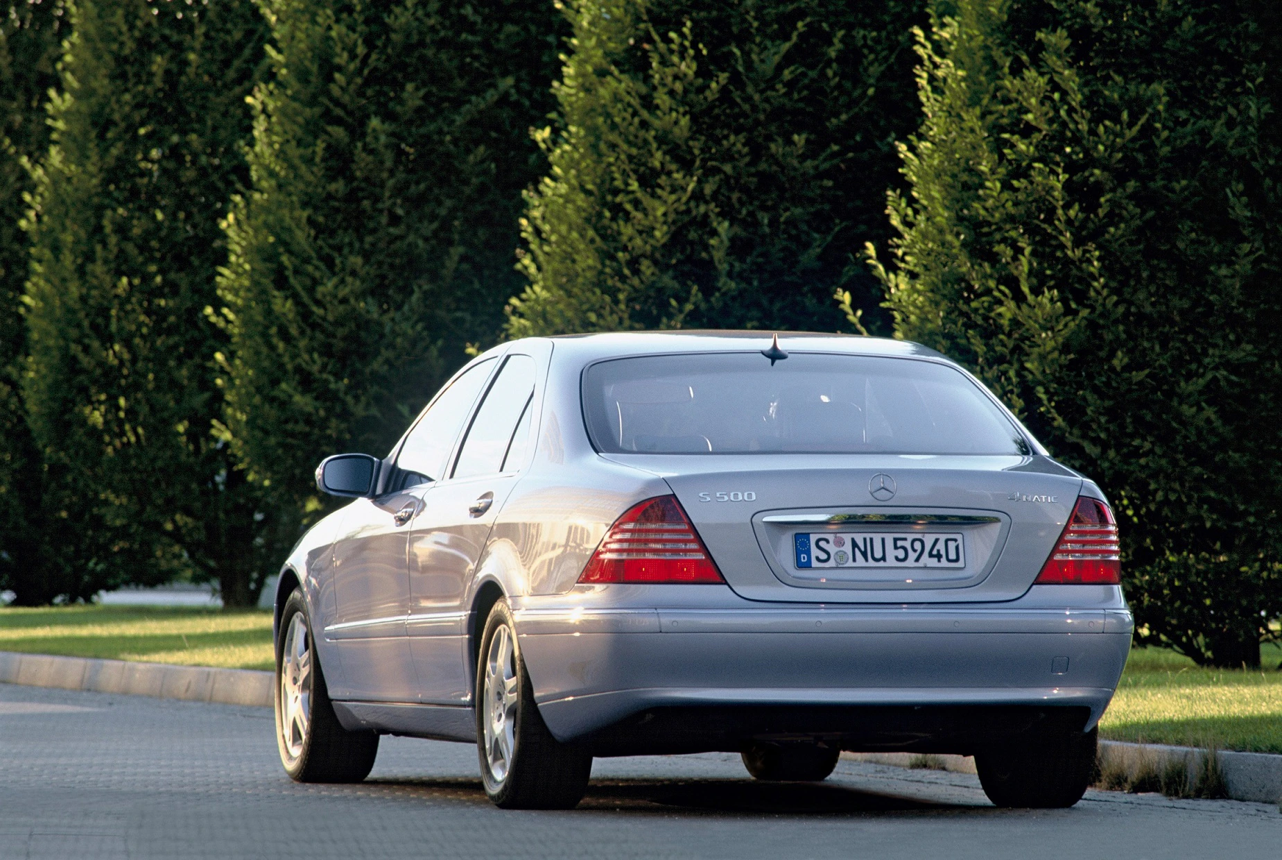 Silberner Mercedes-Benz S-Klasse parkt auf gepflasterter Straße, im Hintergrund große, grüne Bäume.