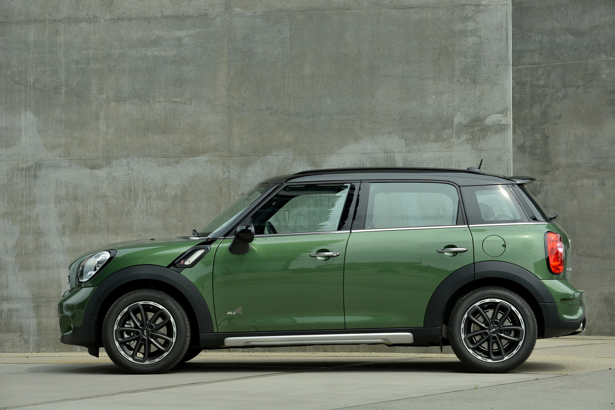 Grüner Mini Cooper SUV steht seitlich vor einer grauen Betonwand auf einem asphaltierten Boden.