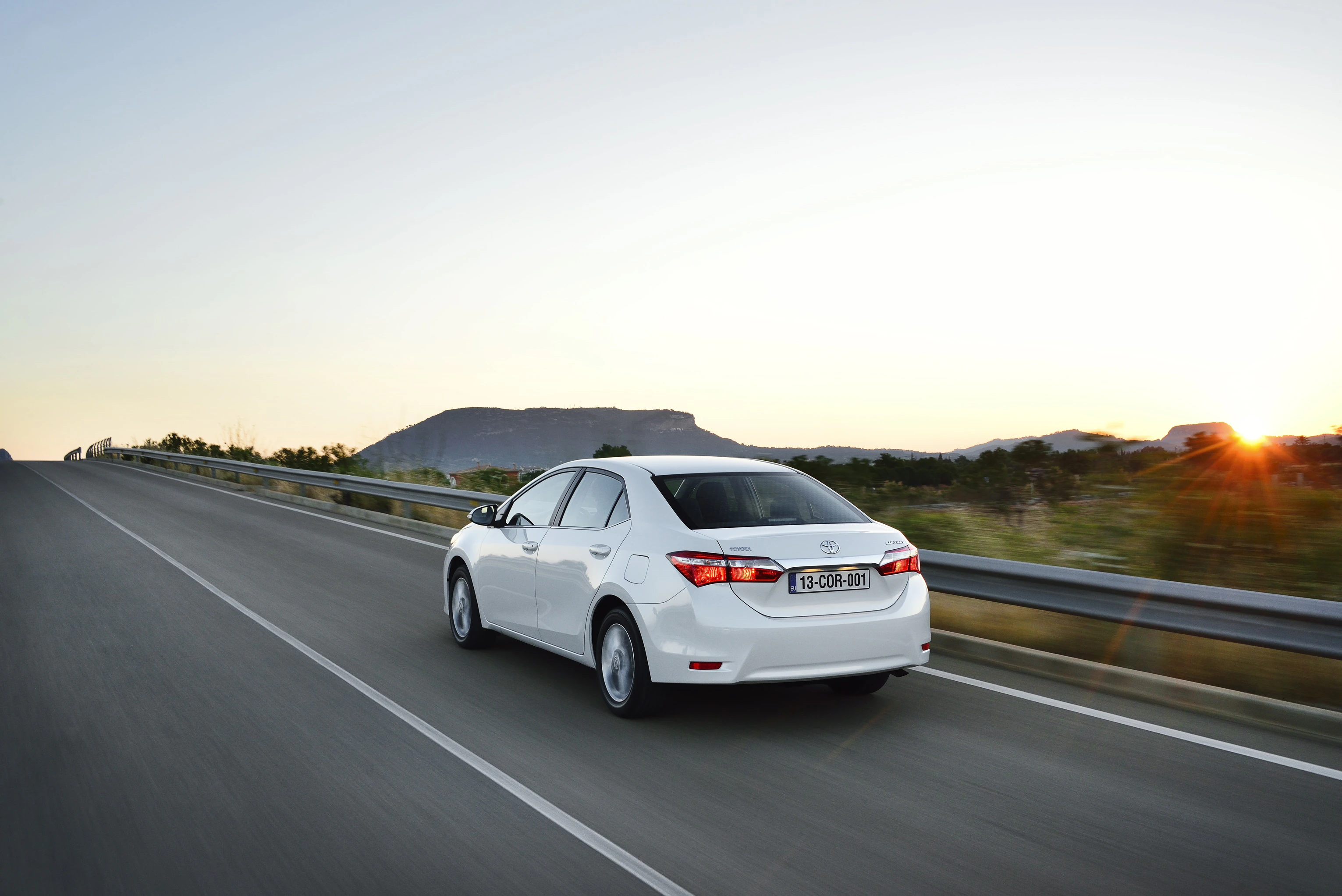 Ein weißes Auto fährt bei Sonnenuntergang auf einer leeren Autobahn, umgeben von Hügeln im Hintergrund.
