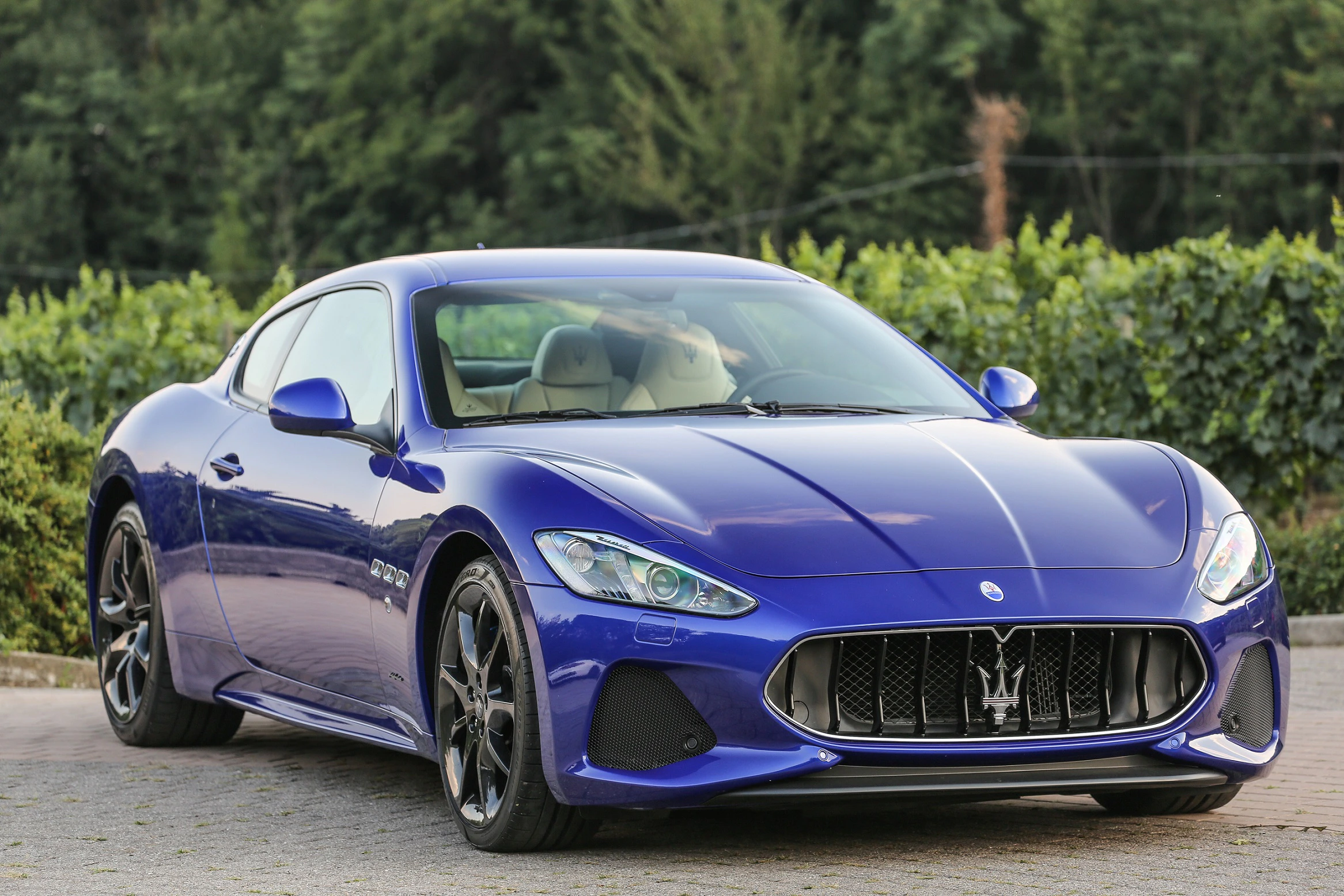 Blauer Sportwagen, Maserati-Modell, geparkt auf einer Straße mit grünem Laub im Hintergrund, vor einem bewölkten Himmel.