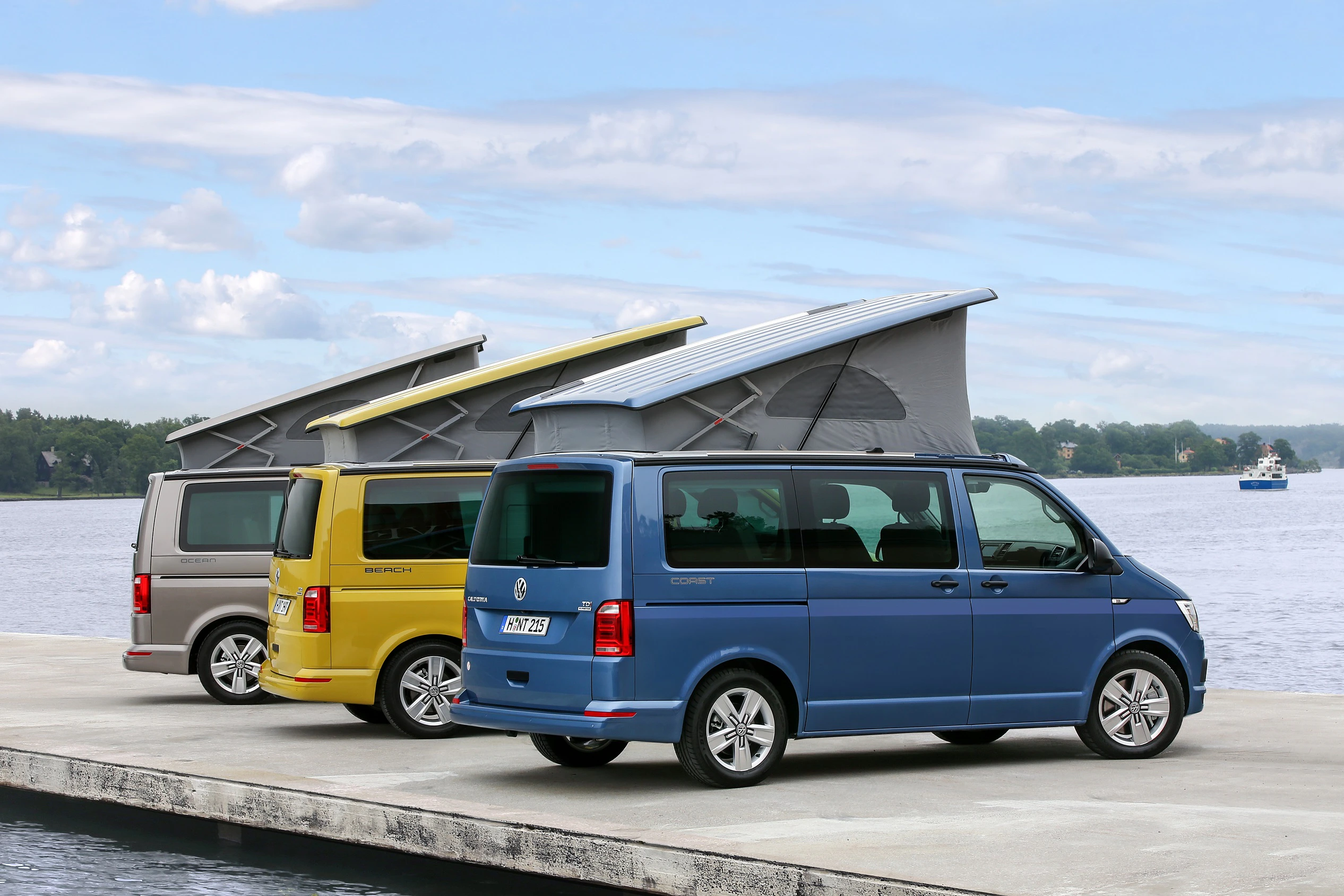 Drei bunte Campingbusse mit aufgeklappten Dachzelten stehen auf einem Steg vor einem See. Im Hintergrund sind Bäume und ein bewölkter Himmel.