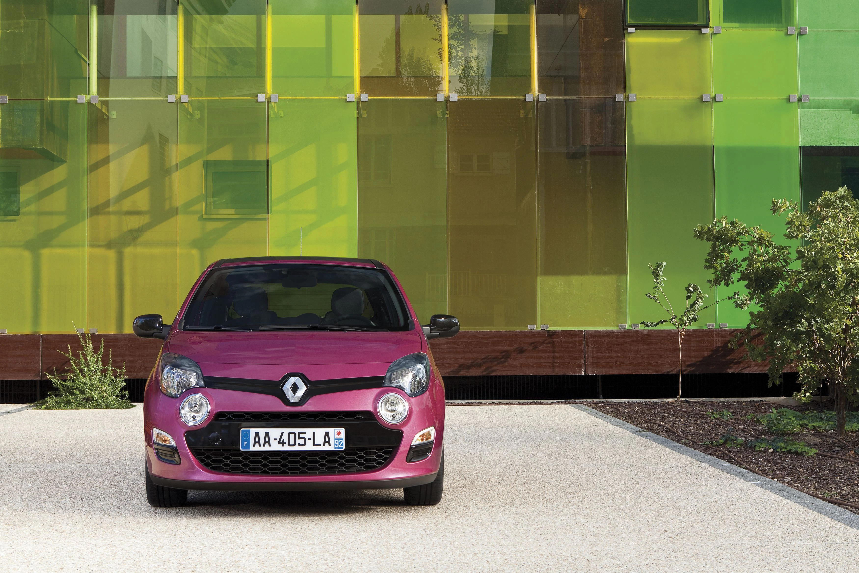 Rosa Renault Twingo parkt vor einem modernen Gebäude mit farbigen Glasfassaden in Gelb- und Grüntönen.
