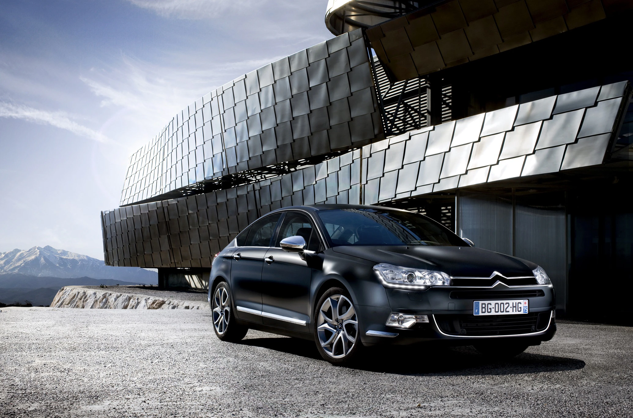 Schwarzer Citroën C5 parkt vor einem modernen Gebäude mit gebogener Glasfassade, im Hintergrund schneebedeckte Berge.
