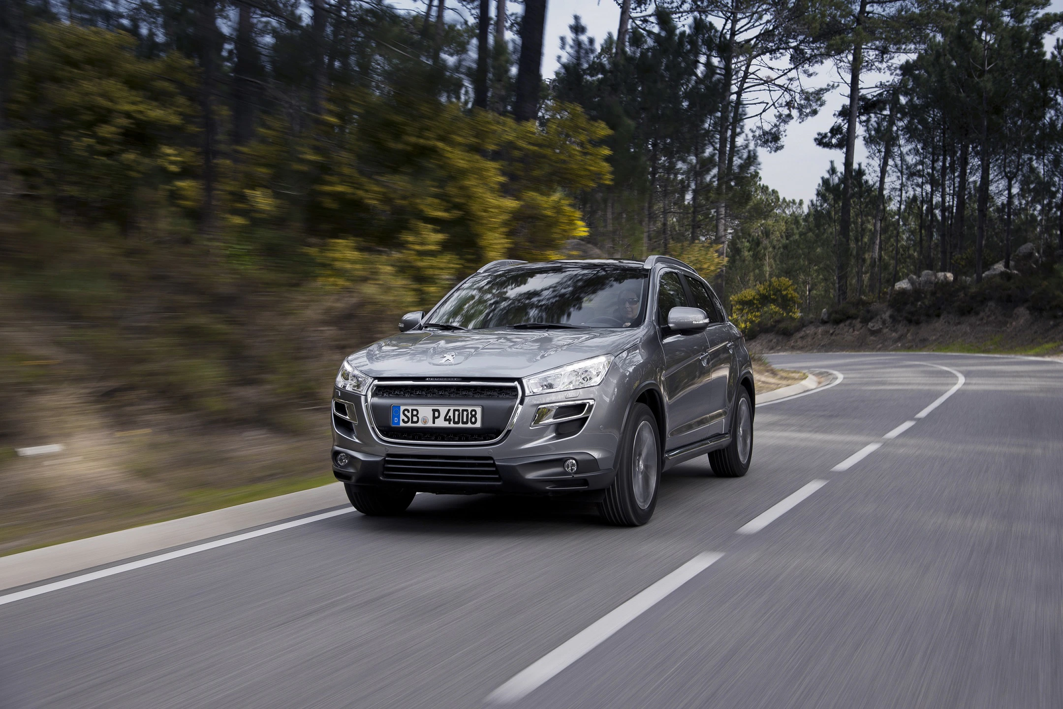 Silberner SUV fährt auf kurviger Landstraße durch waldreiche Landschaft bei Tageslicht.