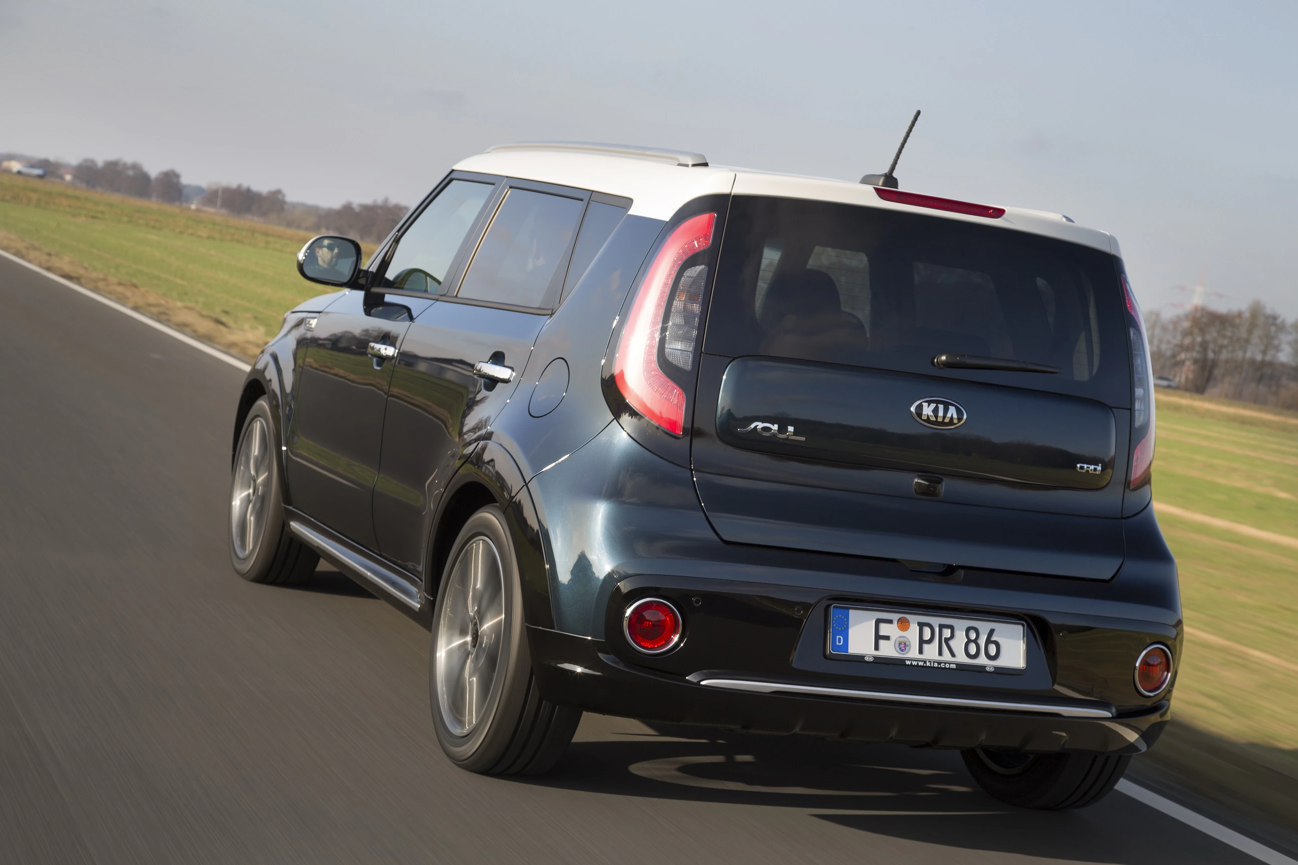 Ein dunkelgrauer Kia Soul fährt auf einer Landstraße, umgeben von grünen Feldern unter einem klaren Himmel.