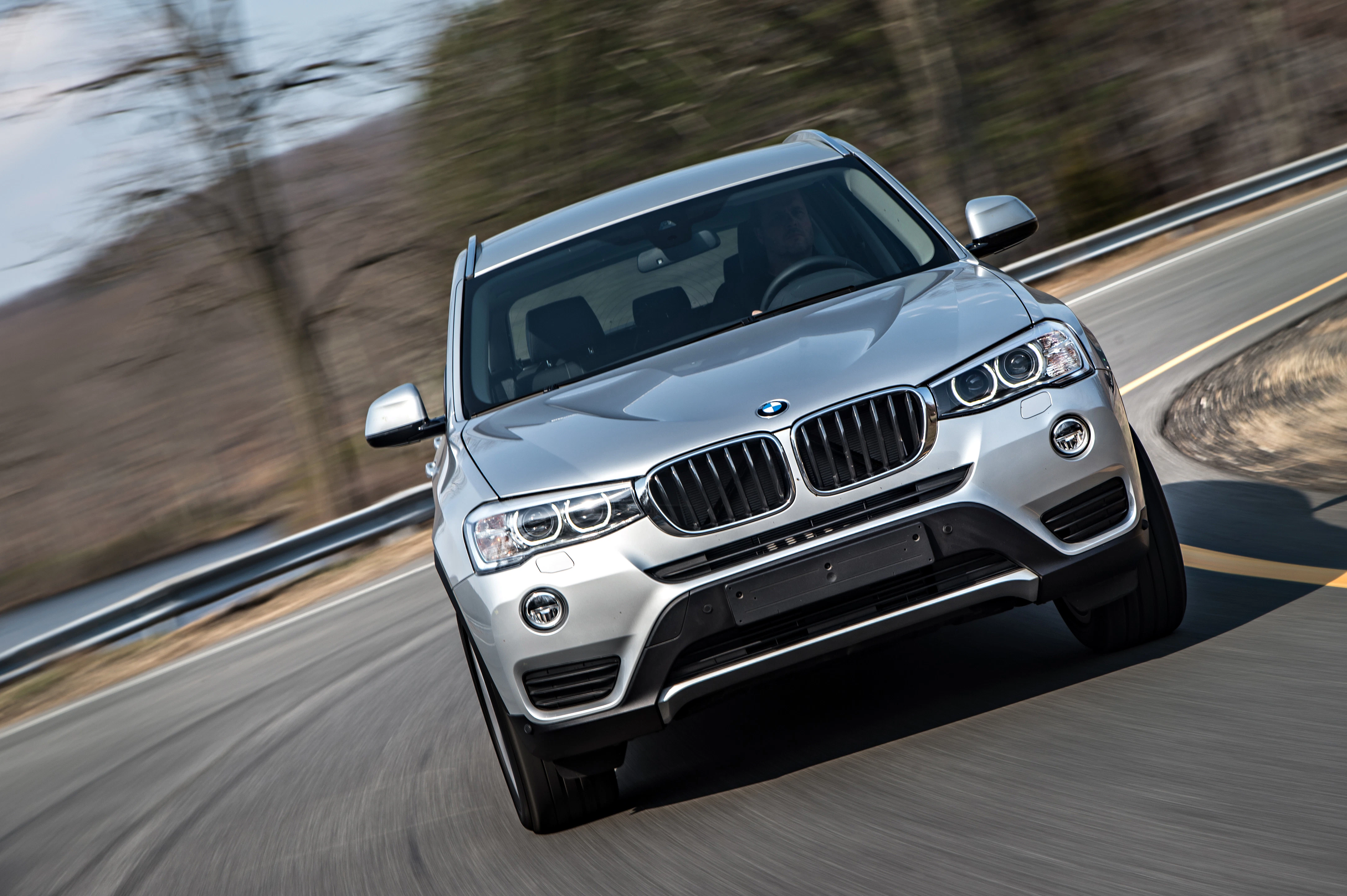 Silberner BMW SUV fährt auf kurviger Landstraße bei bewölktem Himmel, Bäume im Hintergrund, Fahrzeug von vorne gesehen.