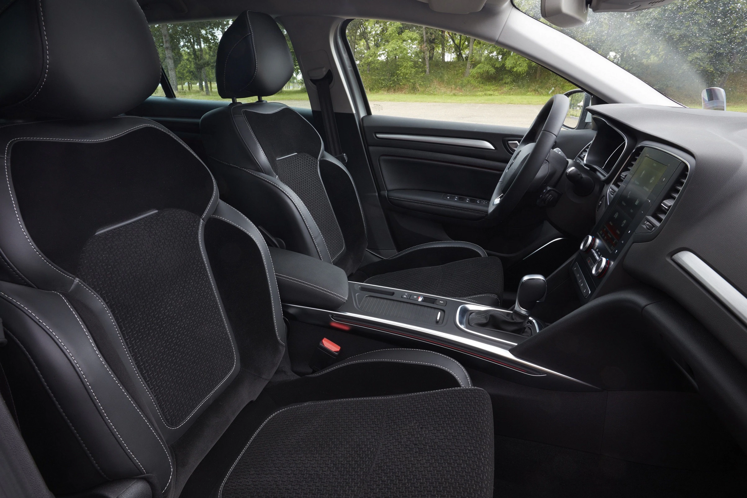 Innenraum eines modernen Autos mit schwarzen Sitzen, Mittelkonsole, Schalthebel und digitalem Display. Fenster zeigen grüne Bäume draußen.