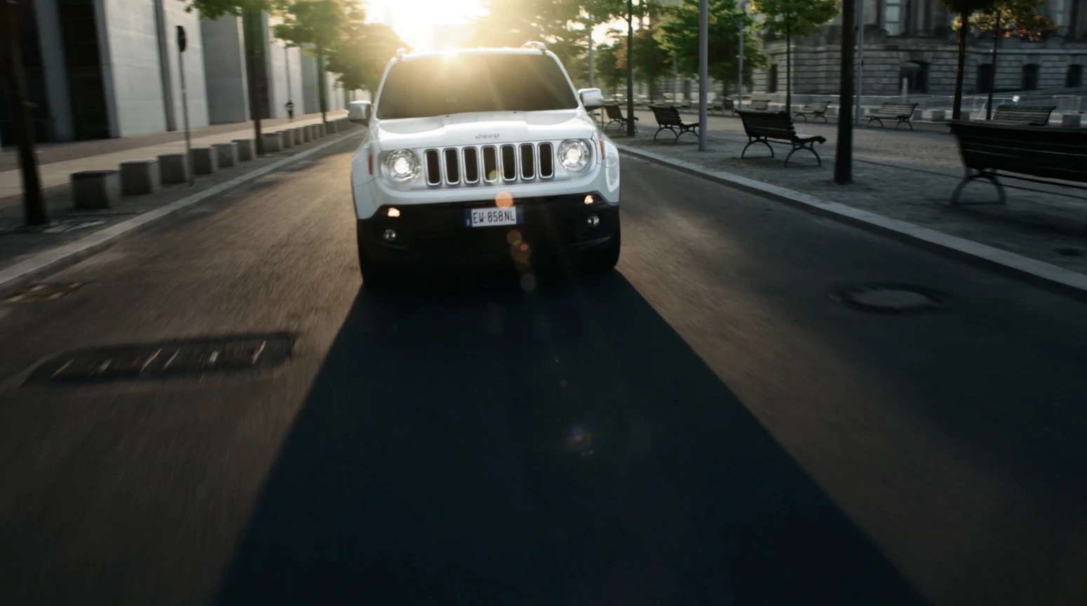 Weißer SUV fährt bei Sonnenuntergang auf leerer, städtischer Straße. Umgeben von Bäumen, Gebäuden und Bänken.
