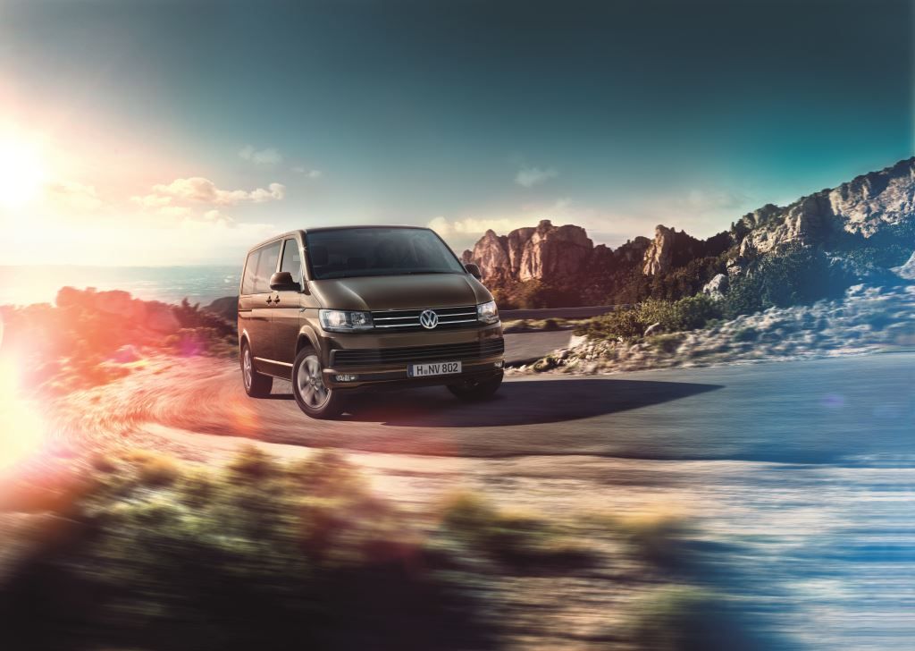 Ein Volkswagen Transporter fährt auf einer kurvigen Bergstraße bei Sonnenuntergang, umgeben von Felsen und dramatischem Himmel.