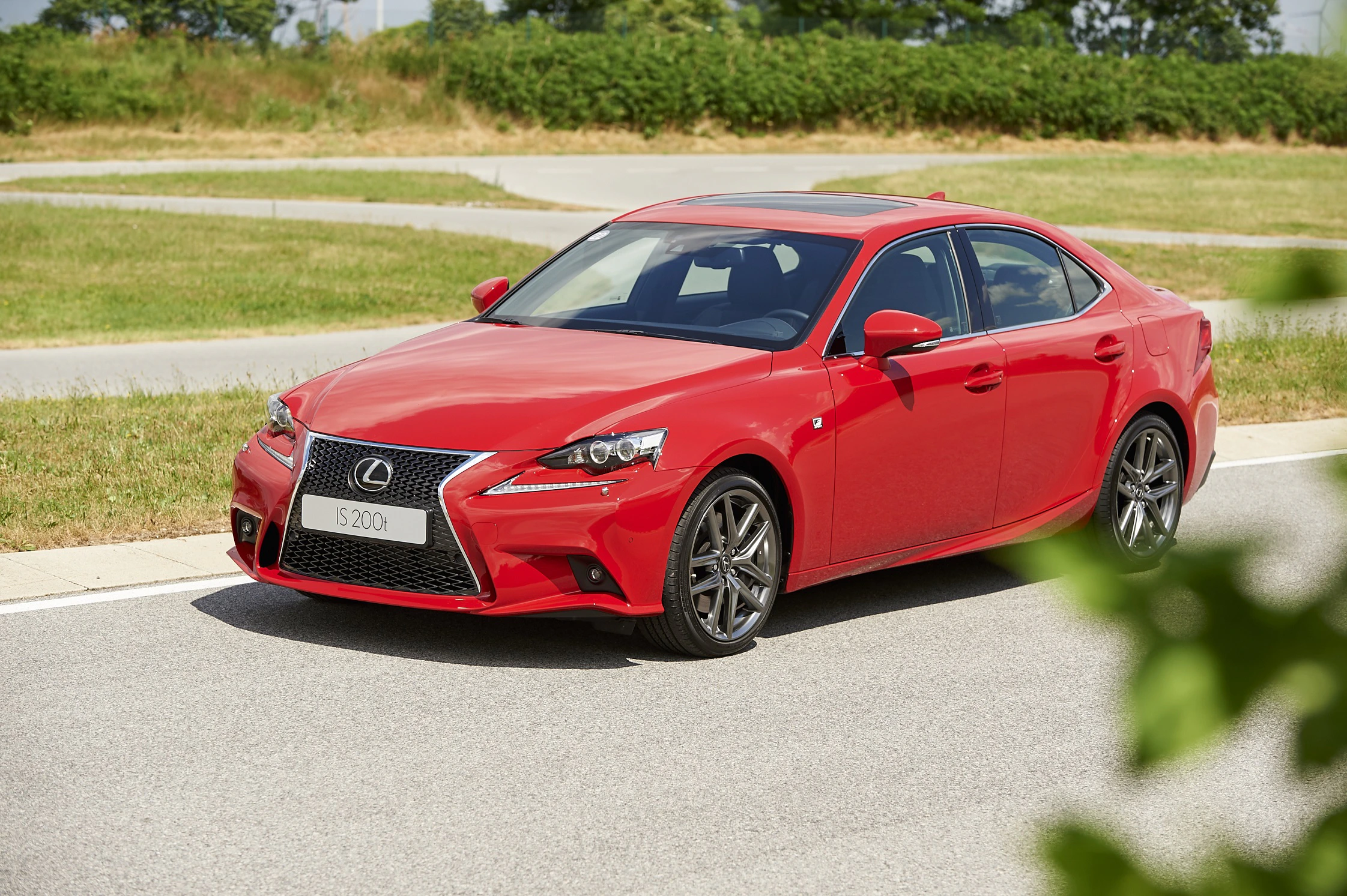 Roter Lexus IS 300h parkt auf einer Straße, umgeben von grünen Büschen und Bäumen im Hintergrund.