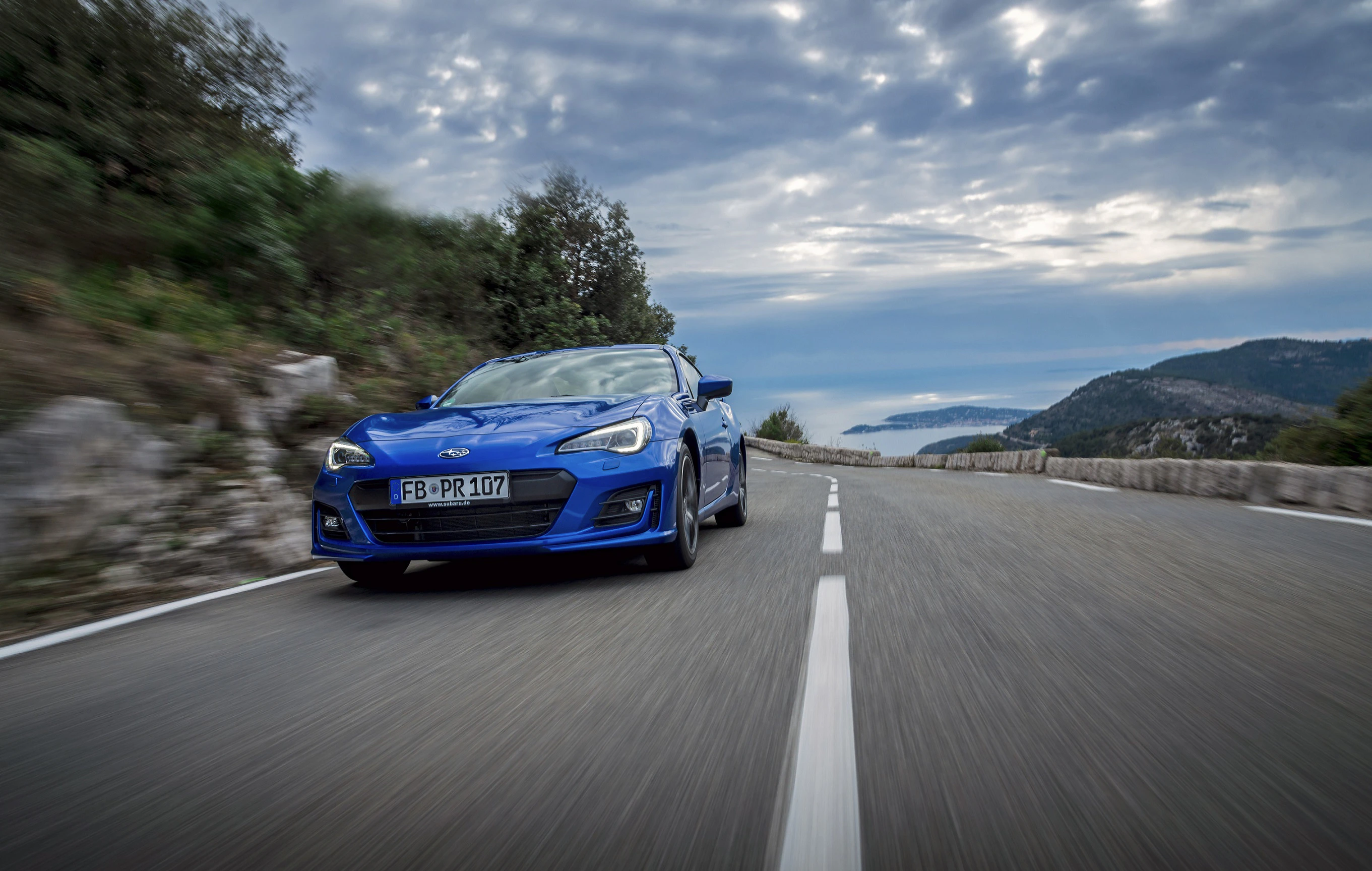 Blauer Sportwagen fährt auf einer kurvenreichen Küstenstraße unter bewölktem Himmel, mit Meerblick im Hintergrund.