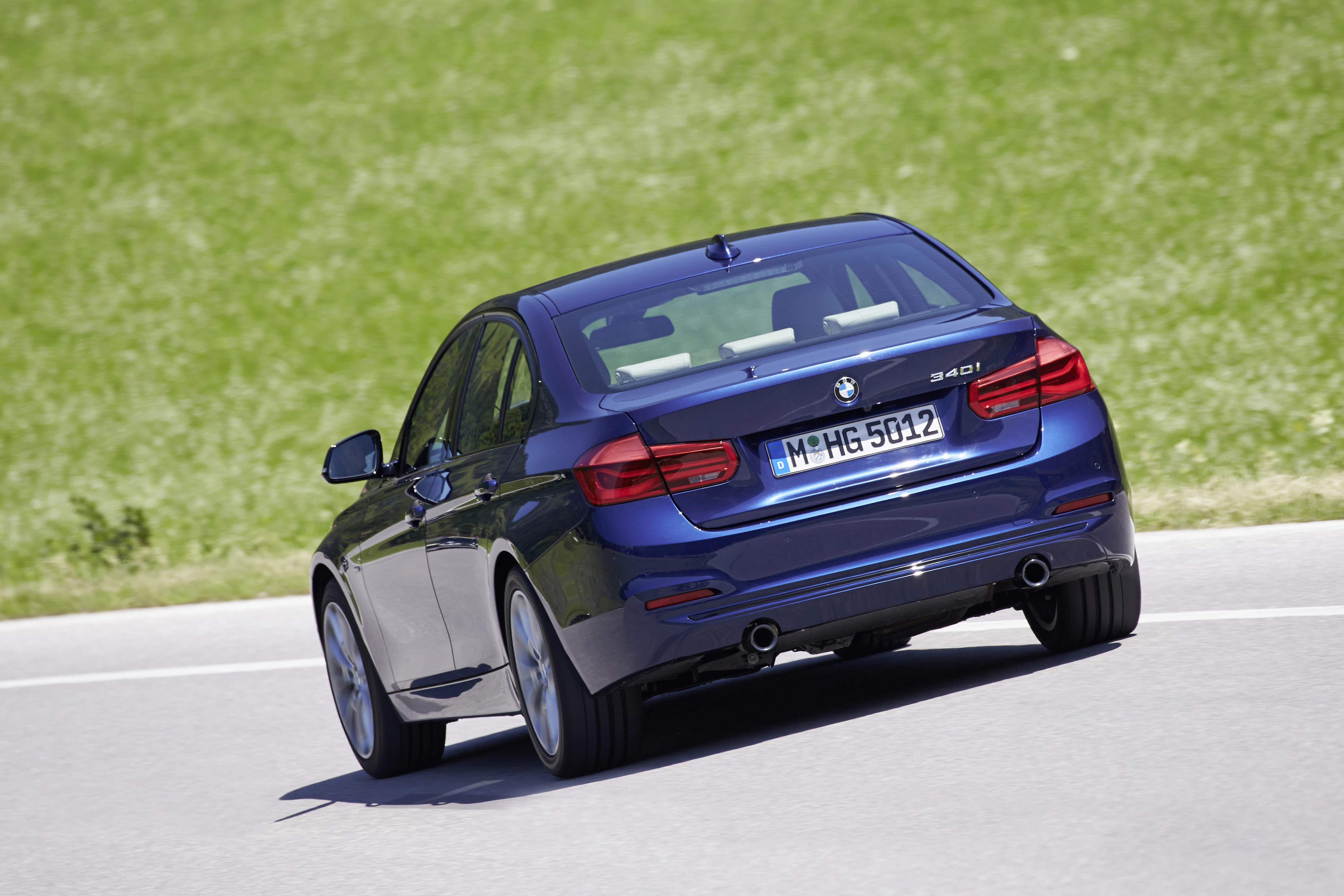 Blauer BMW 3er fährt auf einer Landstraße mit grünem Hintergrund, Rückansicht.