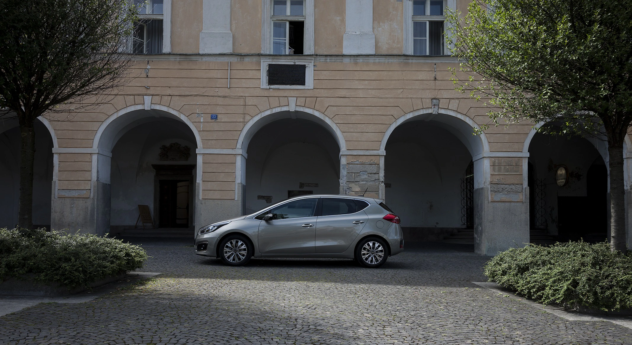 Silbernes Auto vor einem historischen Gebäude mit Arkaden und gepflastertem Hof, umgeben von Bäumen.