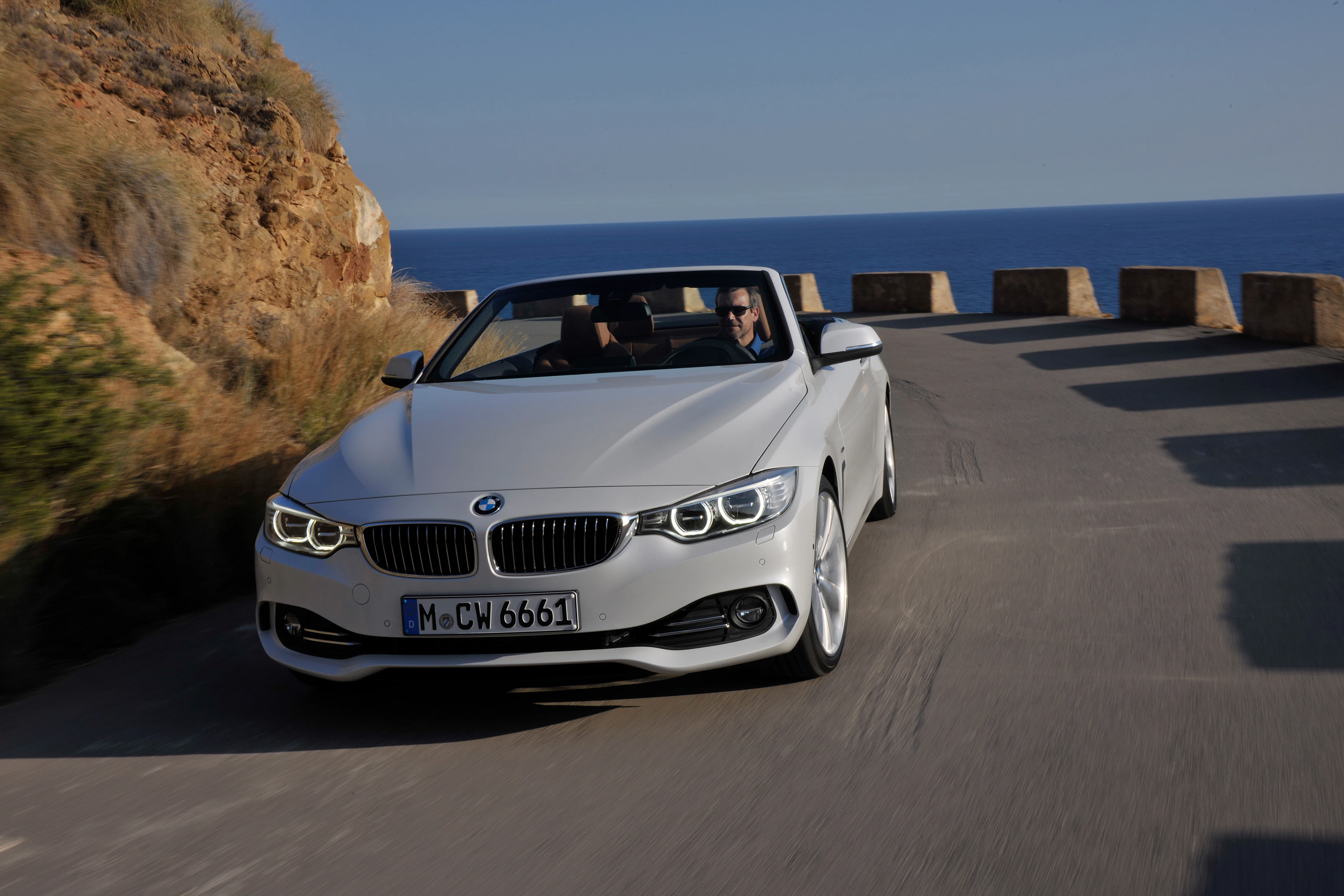 Silbernes BMW-Cabrio fährt auf kurviger Küstenstraße, Fahrer mit Sonnenbrille, Meer im Hintergrund.