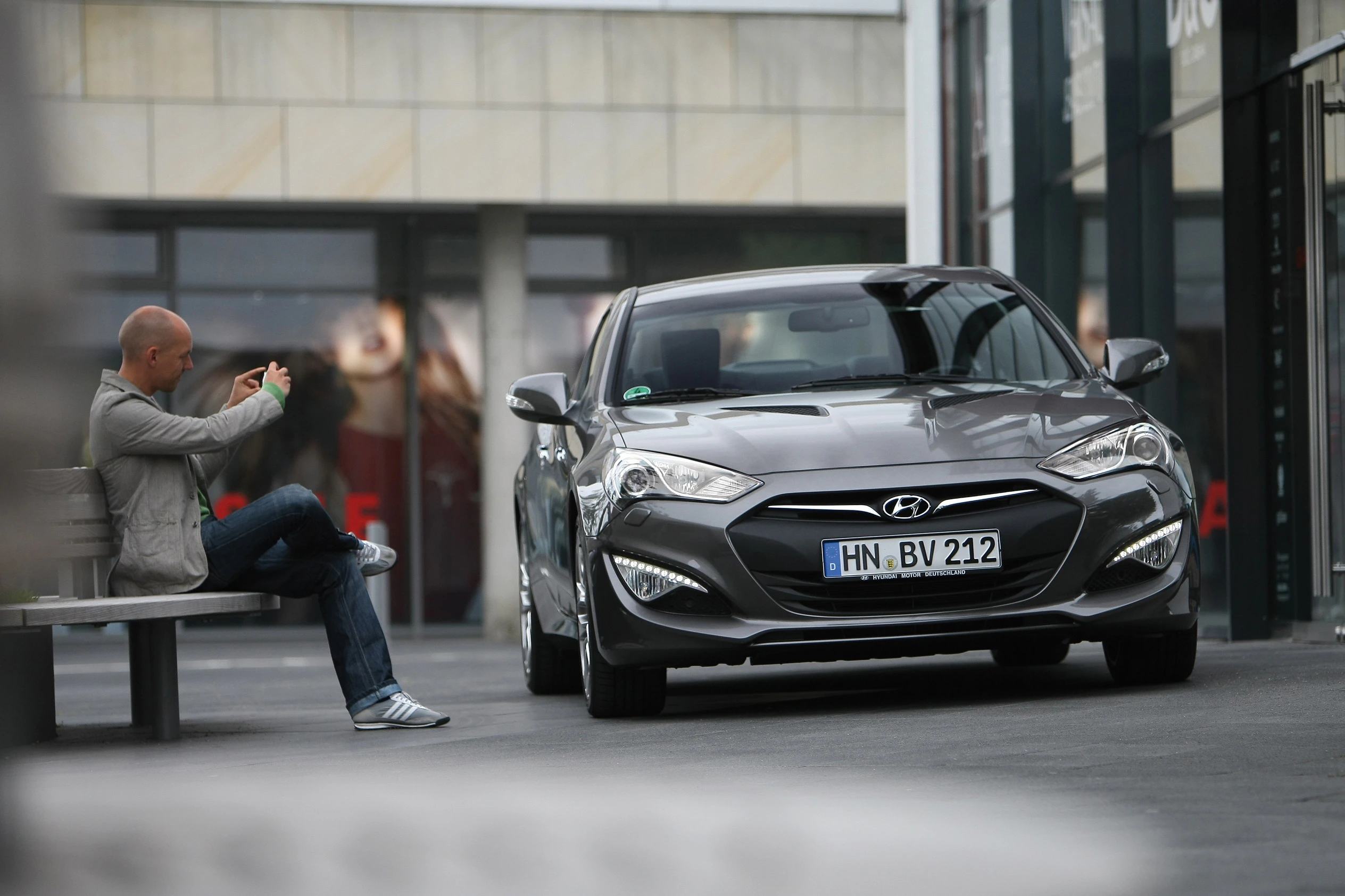 Ein Mann fotografiert auf einer Bank sitzend ein graues Hyundai-Auto, das vor einem modernen Gebäude geparkt ist.