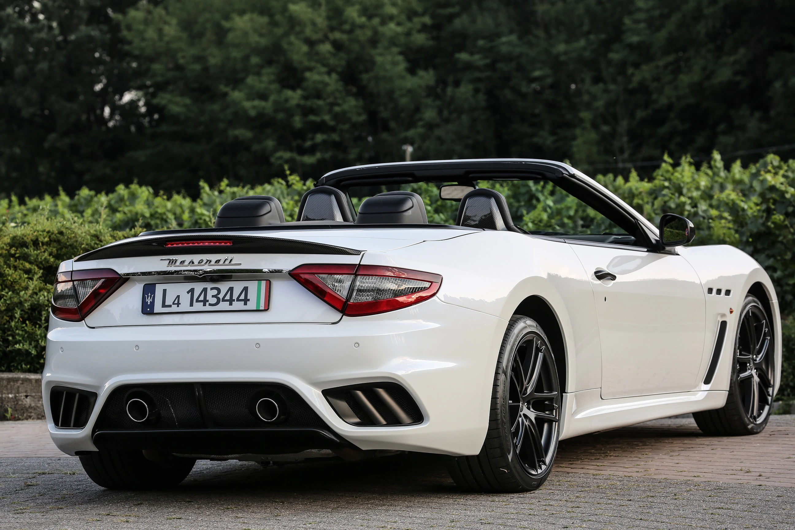 Weißes Maserati-Cabrio mit offenem Verdeck, geparkt vor Bäumen. Das Nummernschild zeigt "44344" und das Auto hat schwarze Felgen.