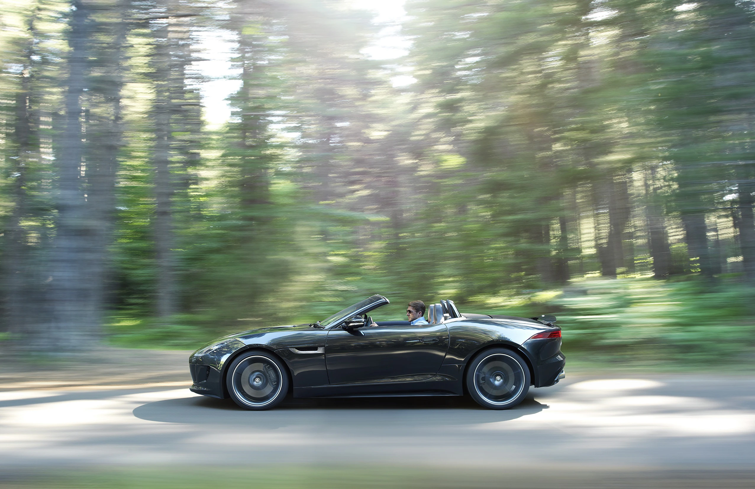 Schwarzes Cabriolet fährt schnell durch einen Wald mit verschwommenen Bäumen, Fahrer am Steuer, Sonnenlicht strömt durch die Baumkronen.