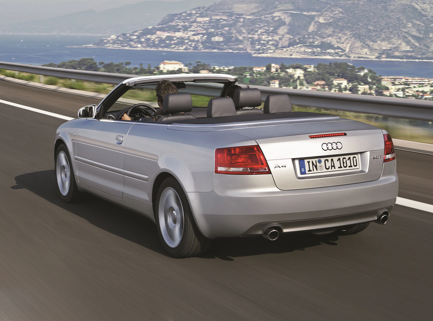 Silbernes Cabriolet fährt auf einer Autobahn, Küstenlandschaft im Hintergrund. Das Auto hat ein geöffnetes Verdeck und ein deutsches Kennzeichen.