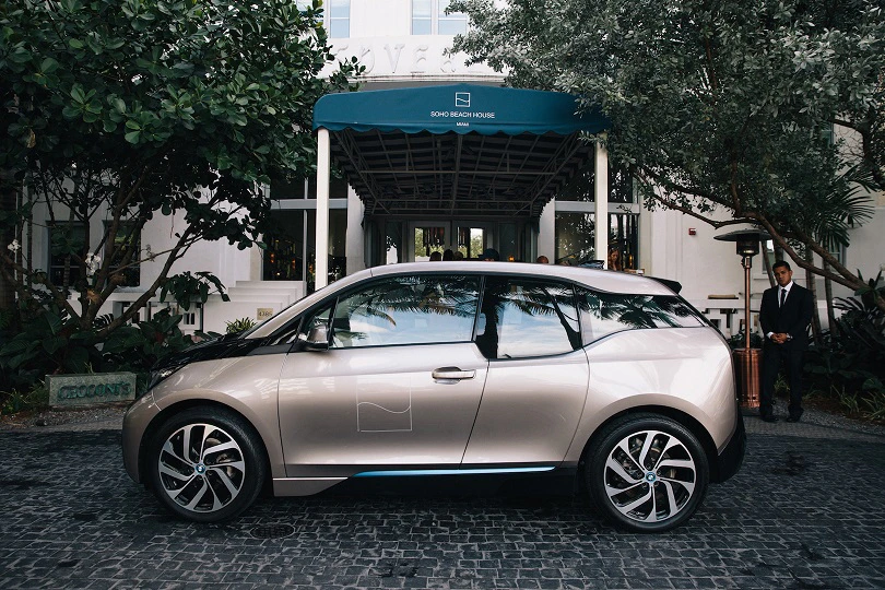 Silberner BMW i3 parkt vor einem eleganten Gebäude mit grünem Dach und umgeben von Bäumen. Eine Person steht daneben.