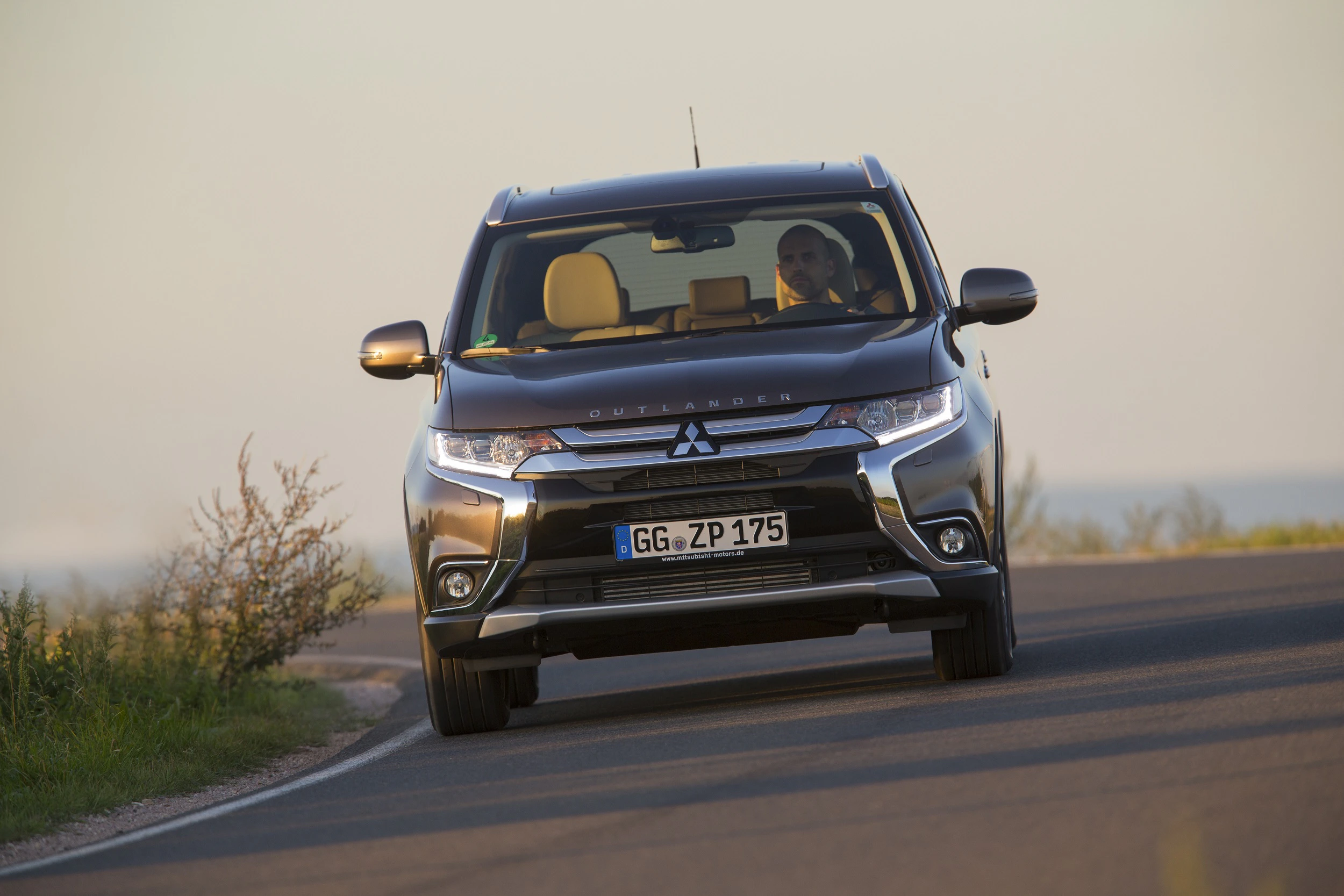 Ein silberner Mitsubishi Outlander fährt auf einer kurvigen Landstraße bei Tageslicht, teilweise von Vegetation umgeben.