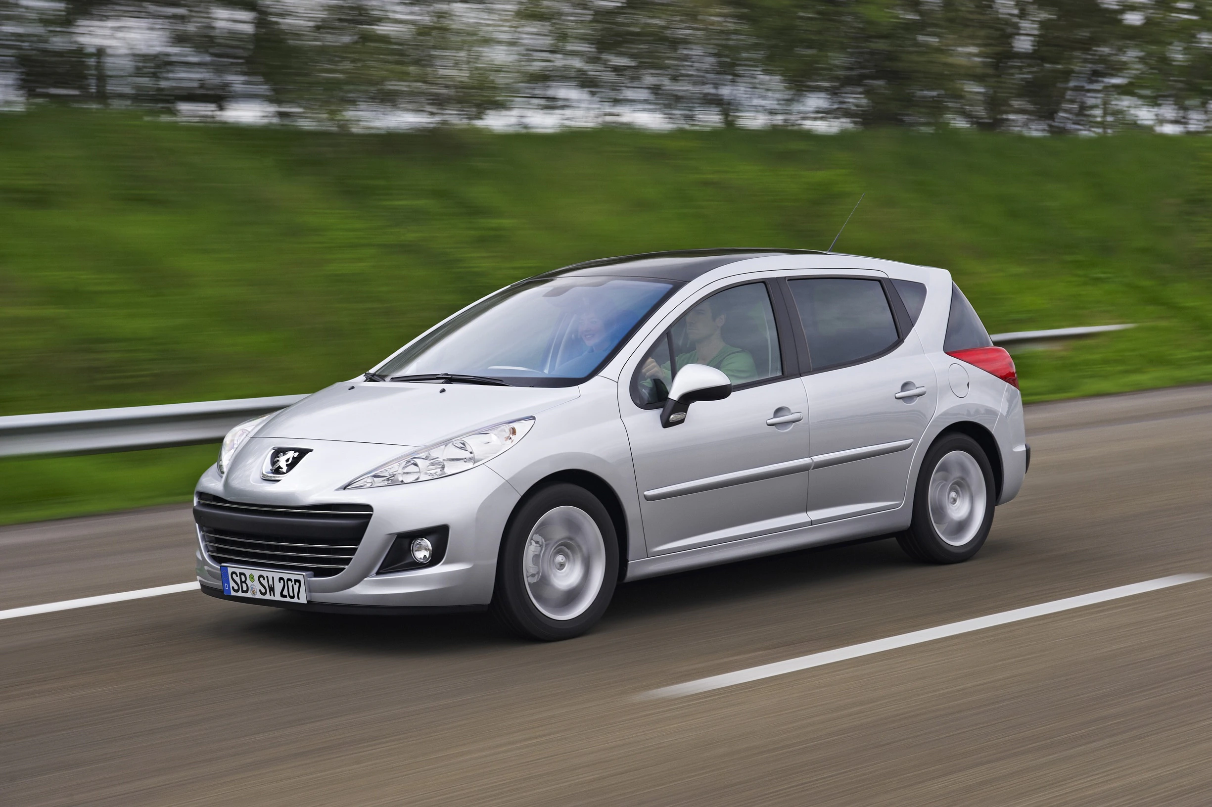 Silberner Peugeot 207 SW fährt auf Straße, grüne Bäume im Hintergrund, leichte Bewegung sichtbar.
