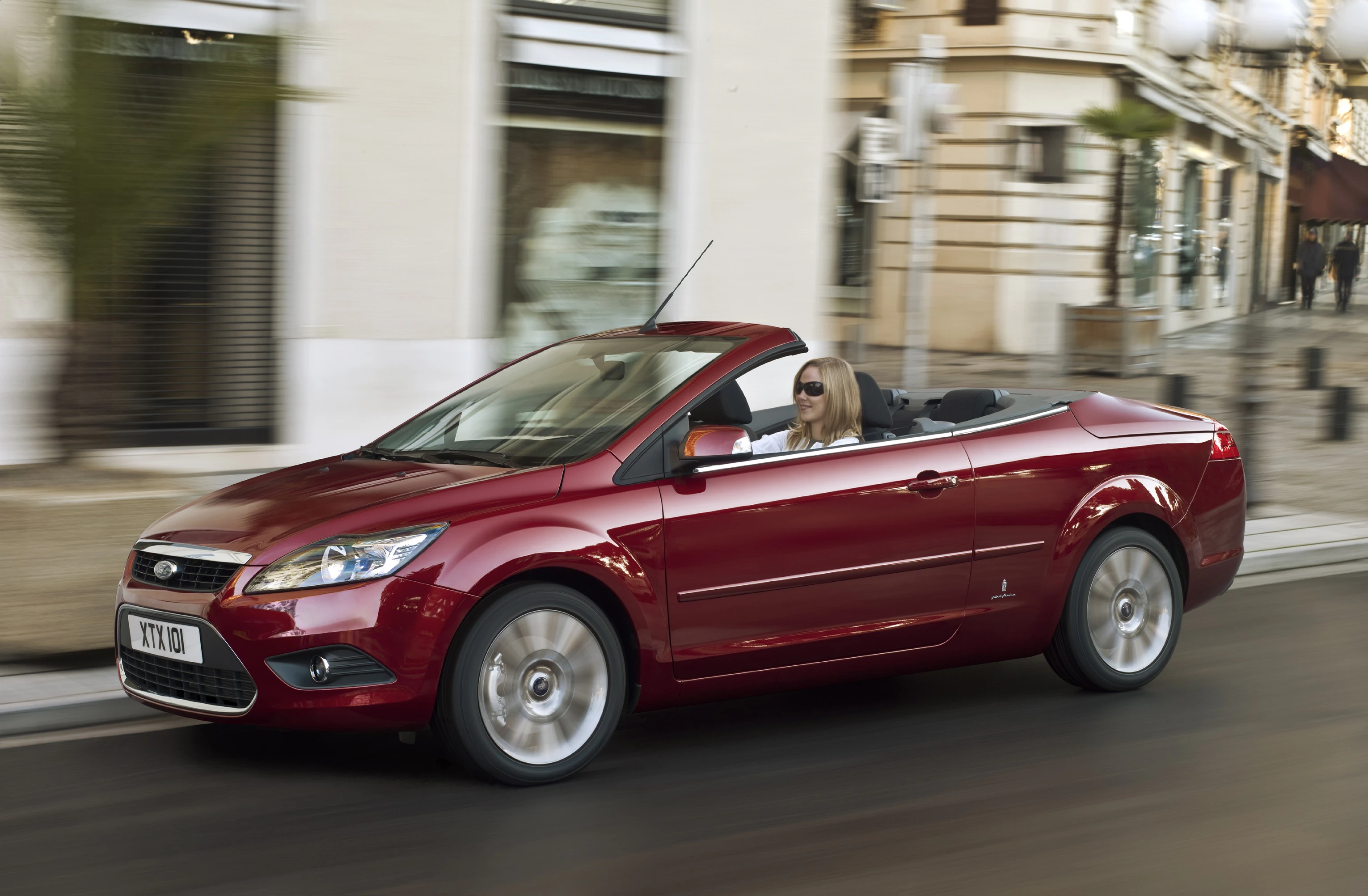Rotes Cabrio mit blonden Fahrer, fahren durch eine städtische Straße bei Tageslicht, von historischen Gebäuden umgeben.