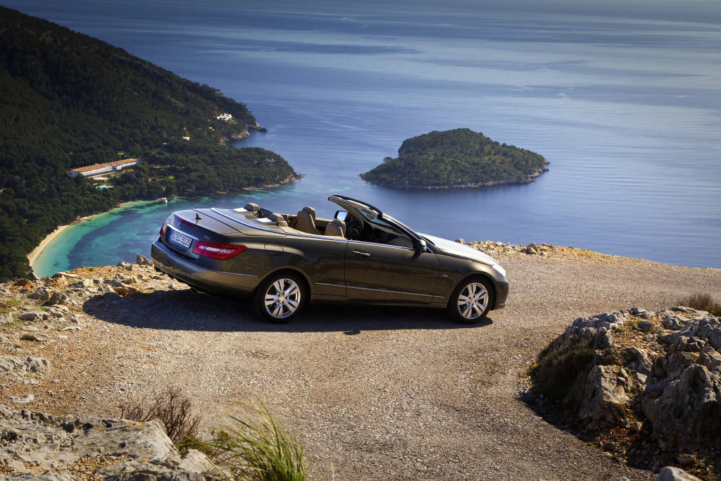Cabrio auf kurviger Küstenstraße mit Blick auf das Meer, umliegende grüne Hügel und eine kleine Insel im Hintergrund.