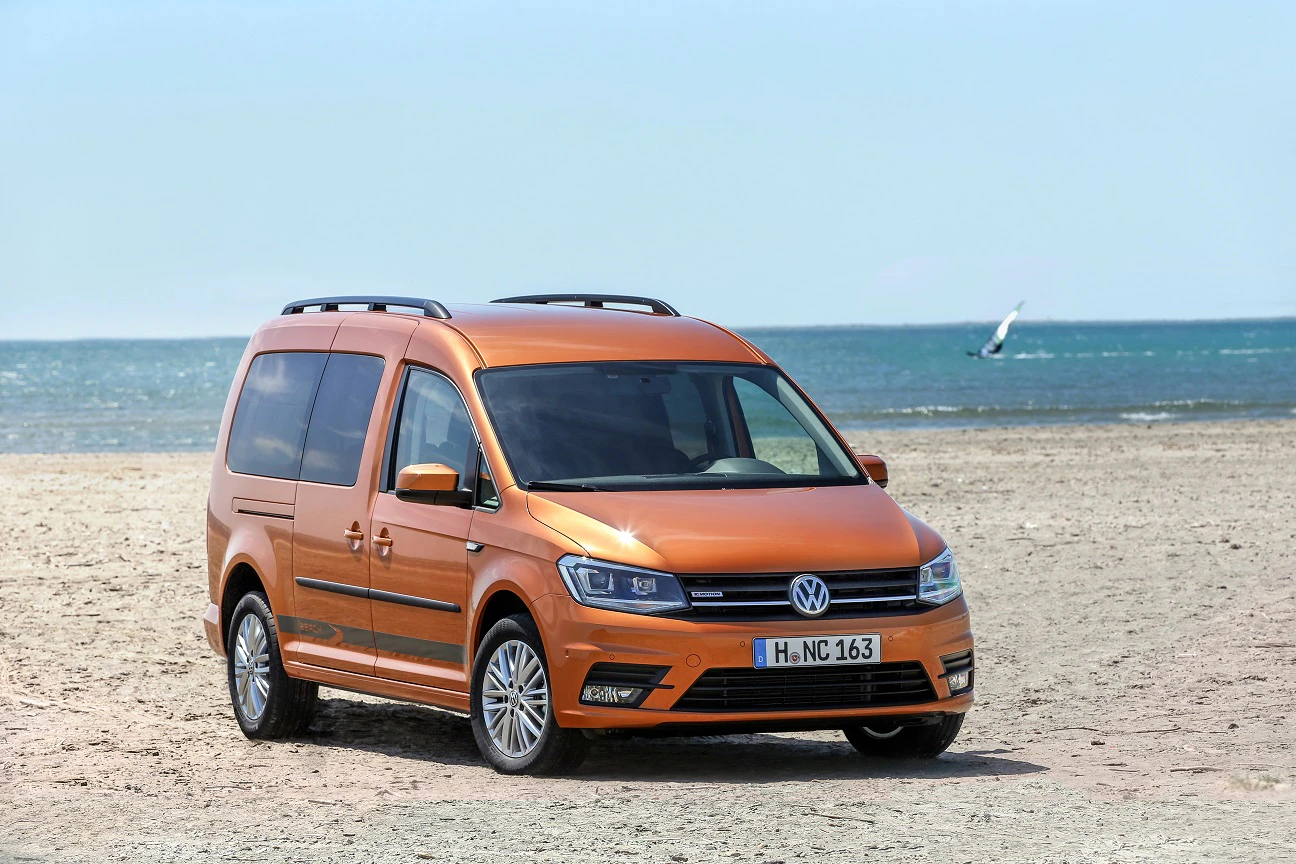 Ein orangefarbener VW Caddy steht am Strand vor blauem Meer und klarem Himmel. Im Hintergrund fährt ein Surfer durch das Wasser.