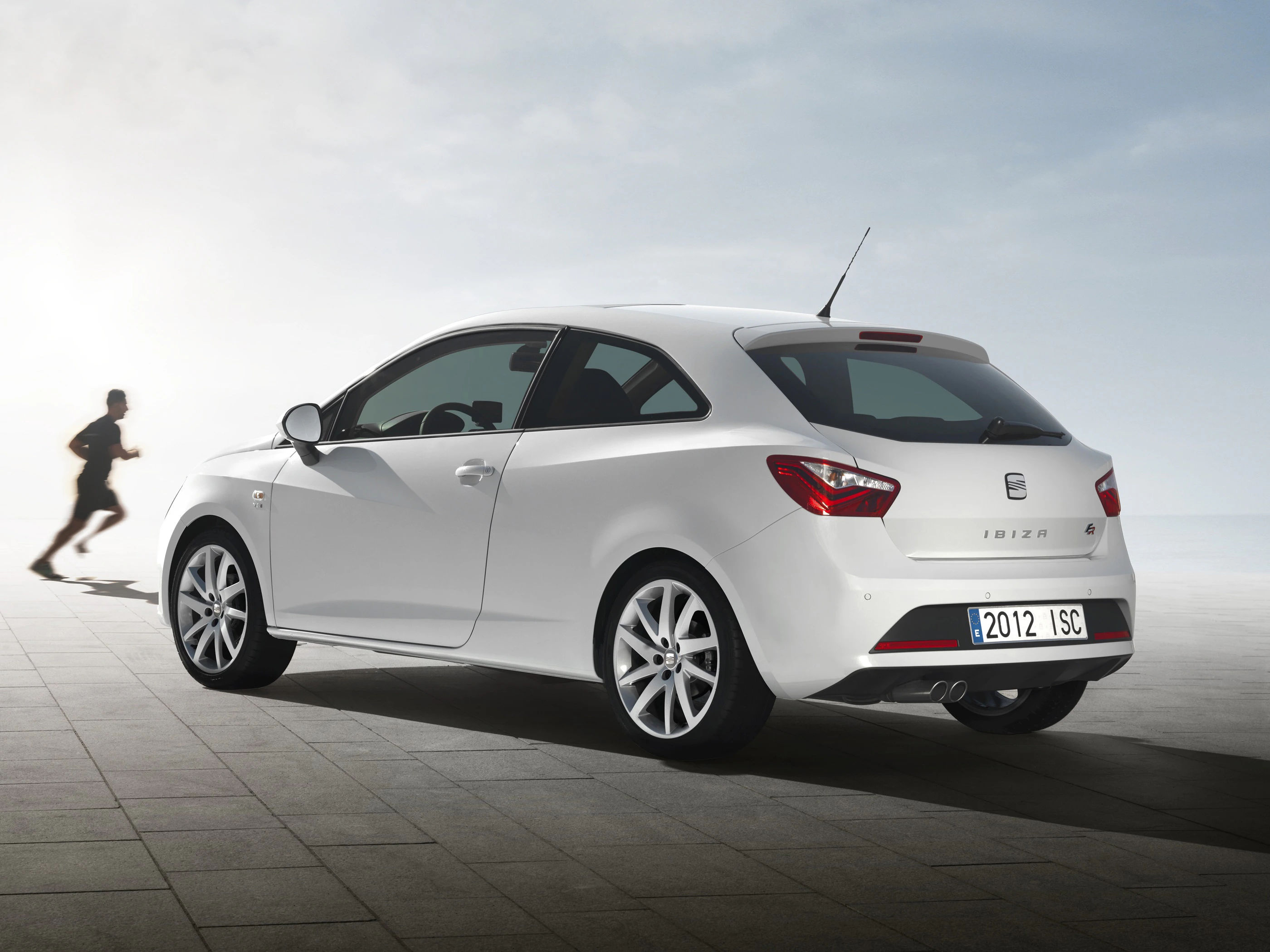 Weißer Seat Ibiza, schräg von hinten fotografiert, auf gepflastertem Platz mit einer laufenden Person im Hintergrund.