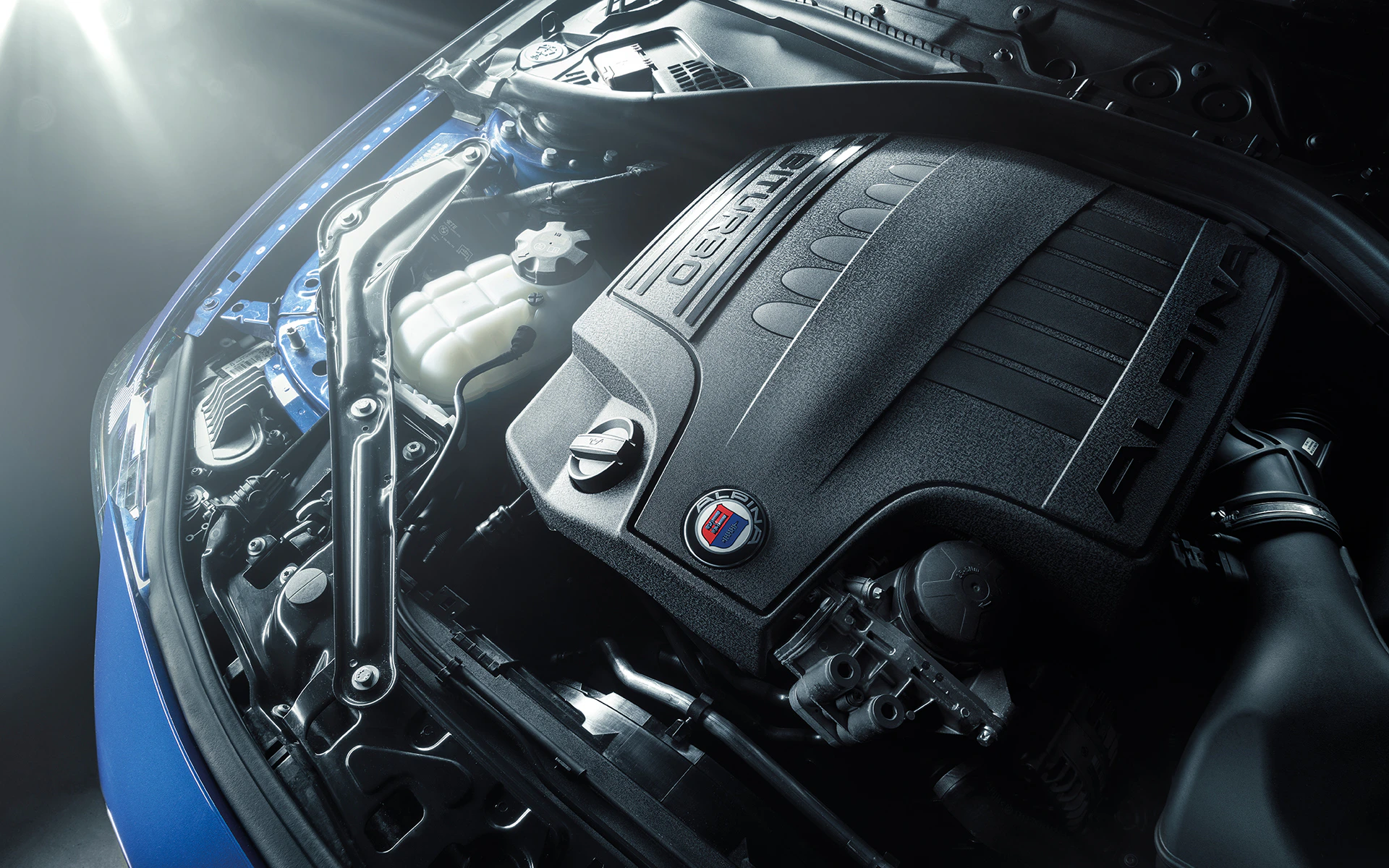 Motorraum eines Autos mit abgedecktem Motor. Zu sehen ist ein Emblem mit der Aufschrift „ALPINA“. Beleuchtung von oben erzeugt Reflexionen.