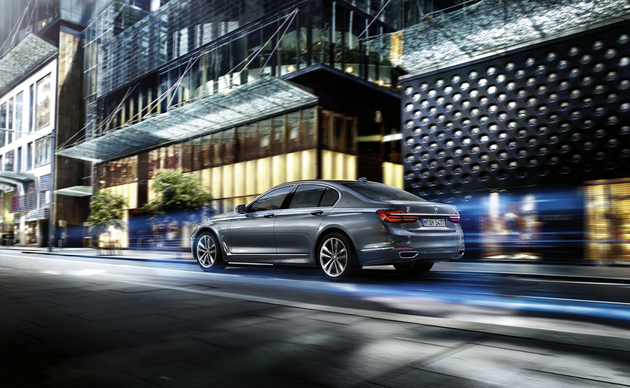 Silbernes Auto fährt dynamisch auf einer städtischen Straße, moderne Gebäude im Hintergrund. Dynamische Lichteffekte erzeugen Geschwindigkeitseindruck.