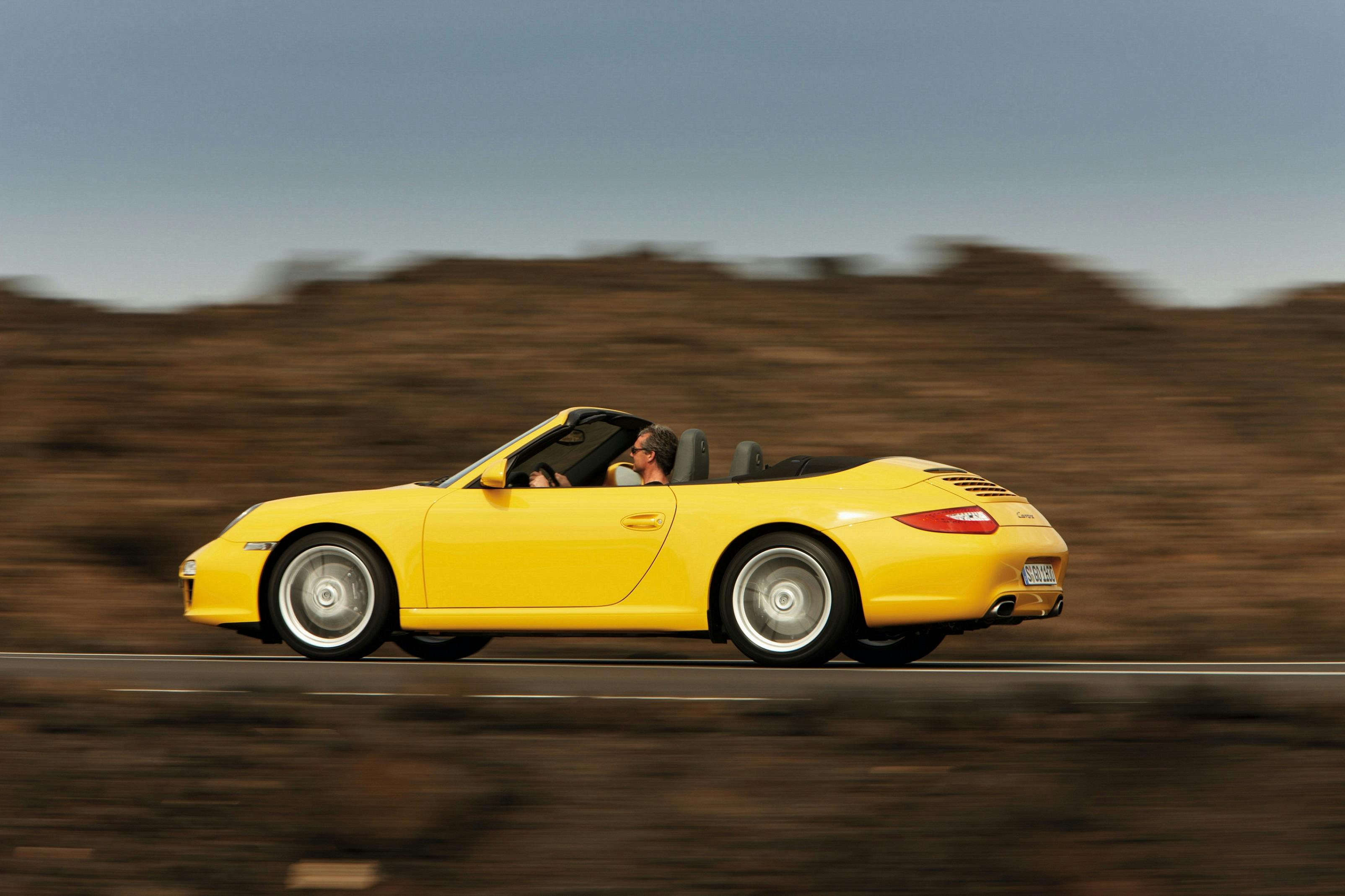 Gelber Sportwagen-Cabrio fährt auf offener Straße, Fahrer am Steuer, verschwommener Hintergrund mit dunklem Himmel.