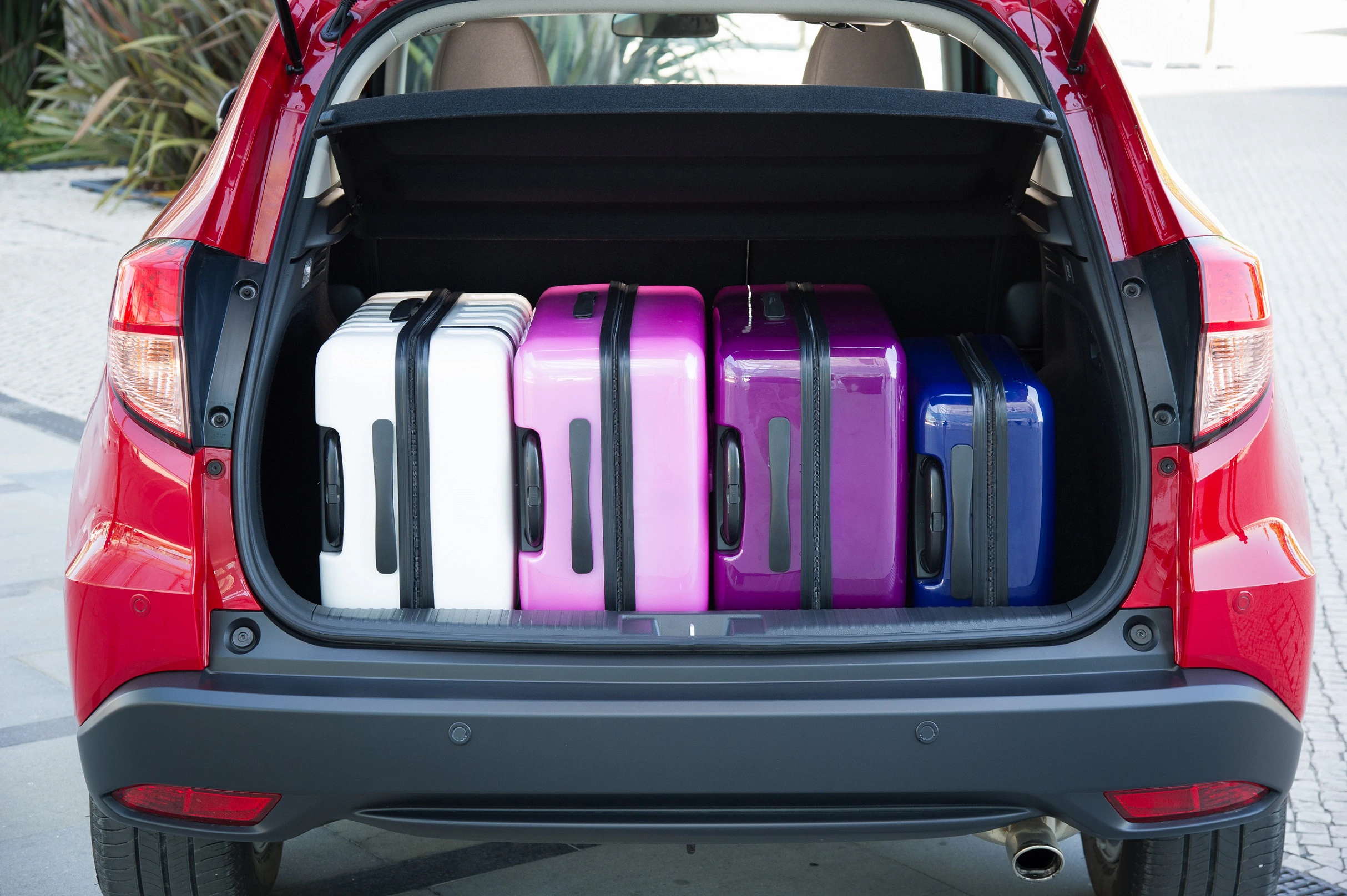 Roter Auto-Kofferraum mit vier farbigen Hartschalenkoffern: weiß, pink, lila und blau. Parken auf gepflastertem Außengelände.