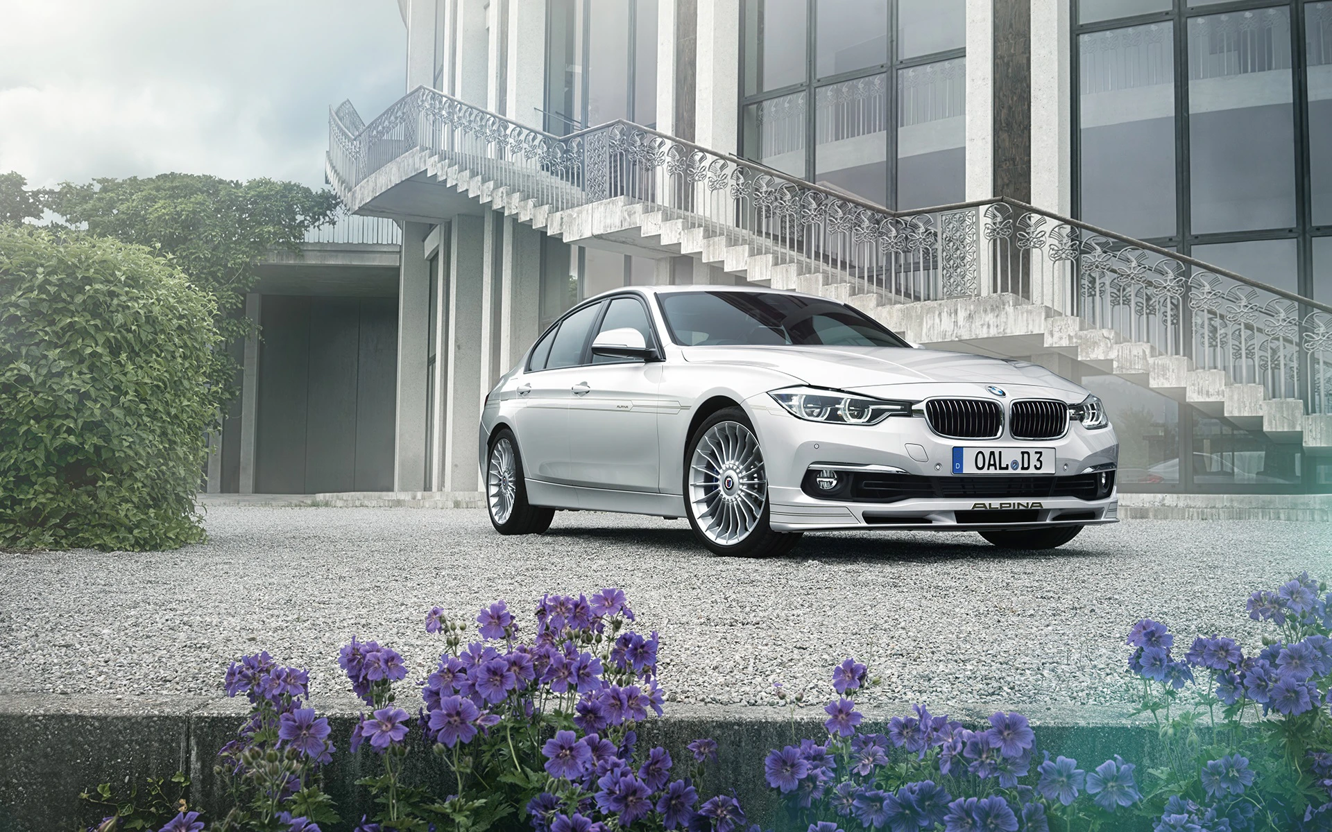 Silberner BMW vor einem modernen Gebäude mit Treppen. Lila Blumen im Vordergrund. Elegantes Design, sonniger Tag.