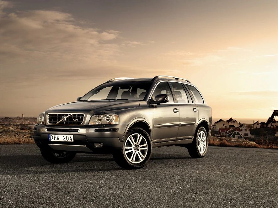 SUV auf leerer Straße bei Sonnenuntergang, umgeben von Landschaft und Häusern im Hintergrund.