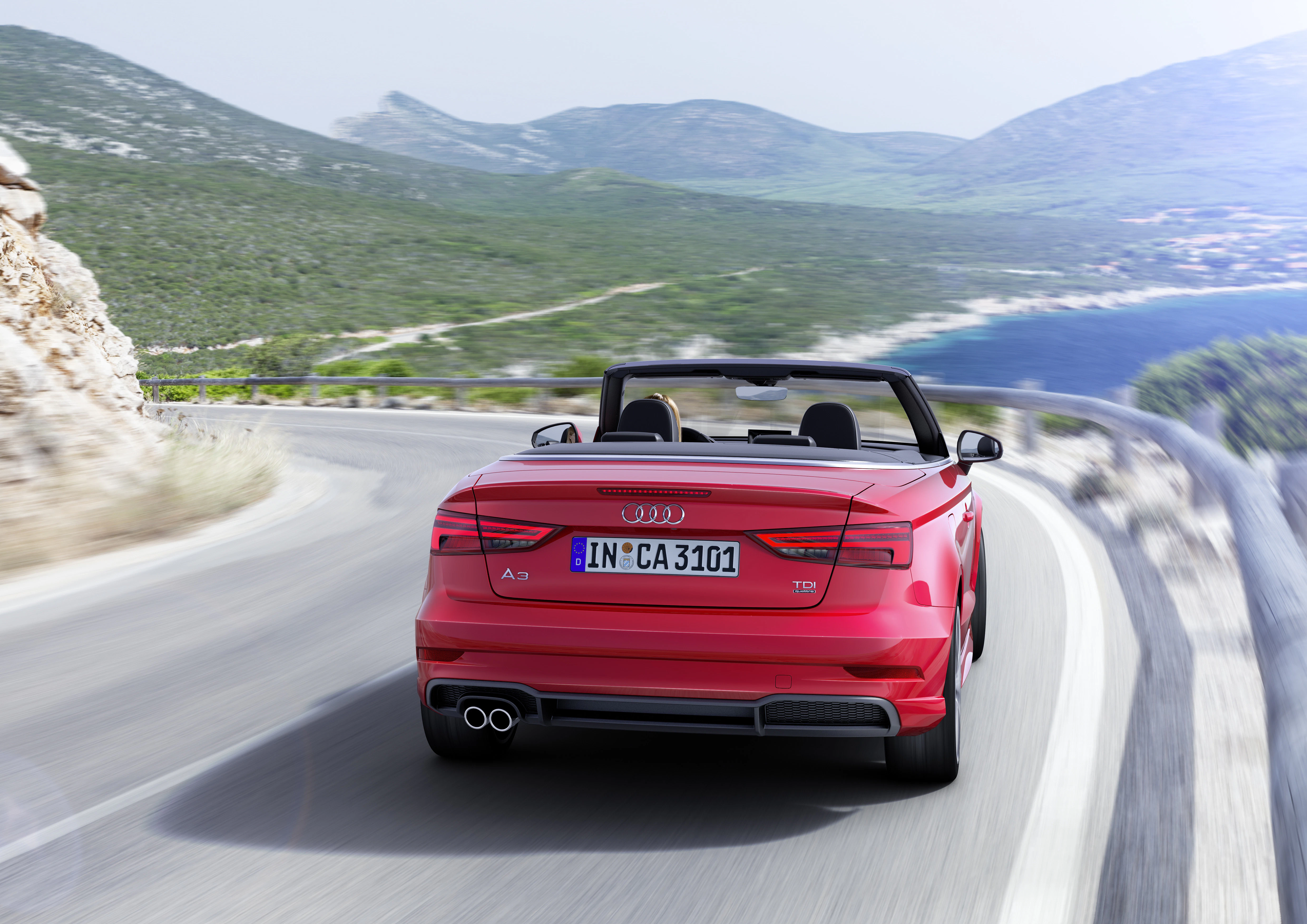 Roter Audi Cabrio fährt auf kurviger Küstenstraße mit Ausblick auf Meer und Berge im Hintergrund.