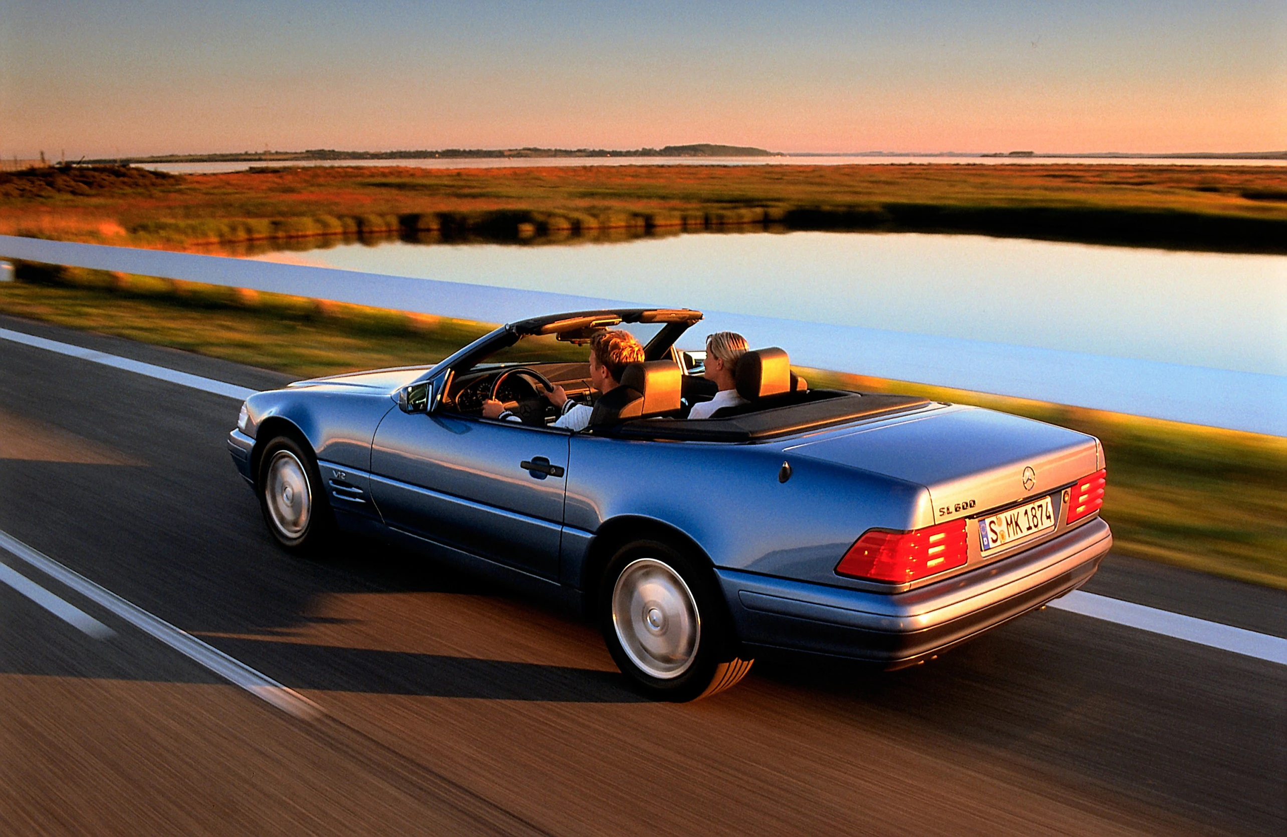 Blaues Cabriolet fährt bei Sonnenuntergang auf einer Landstraße entlang eines Flusses; zwei Personen genießen die Fahrt.