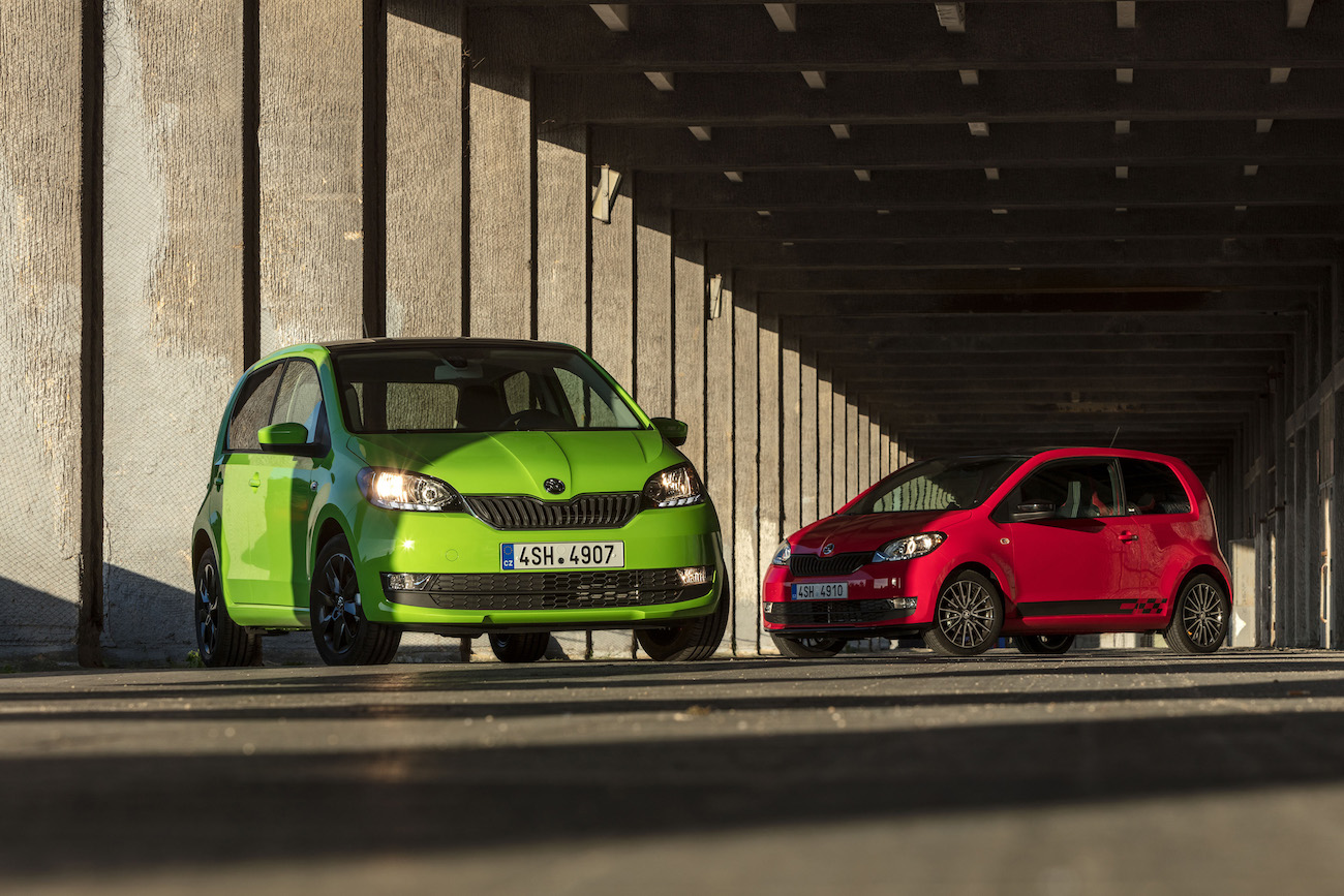 Zwei kleine Autos, eines grün und das andere rot, parken in einer sonnendurchfluteten, überdachten Halle mit Säulen.