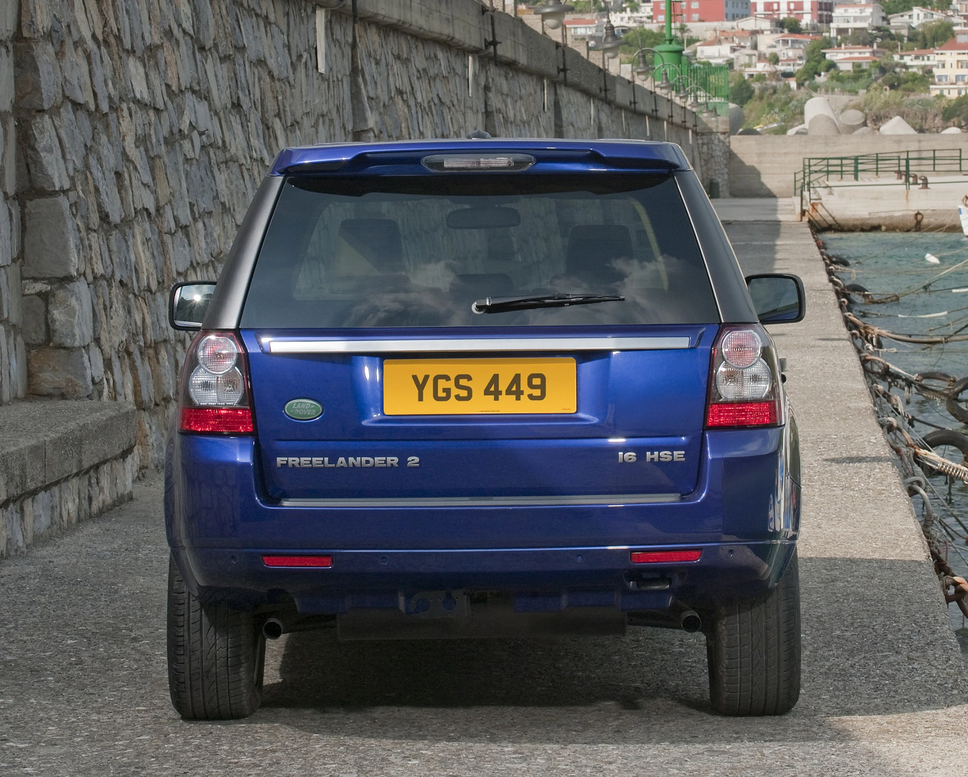 Blauer Land Rover Freelander 2 HSE von hinten, geparkt auf einer Küstenstraße mit steiniger Mauer und Meer im Hintergrund.