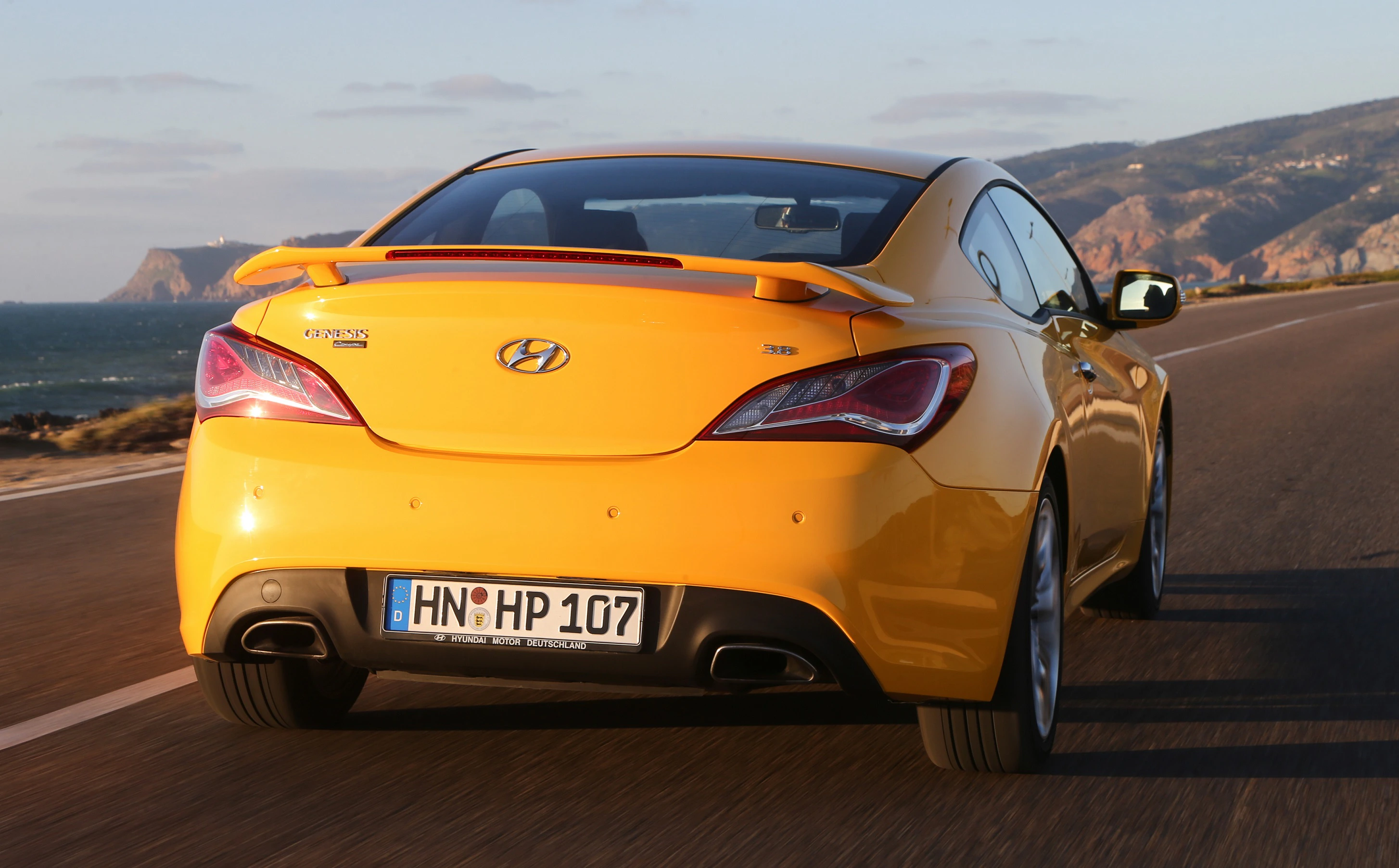 Gelbes Hyundai-Coupé fährt auf einer Küstenstraße bei Sonnenuntergang. Meer und Hügel sind im Hintergrund zu sehen.