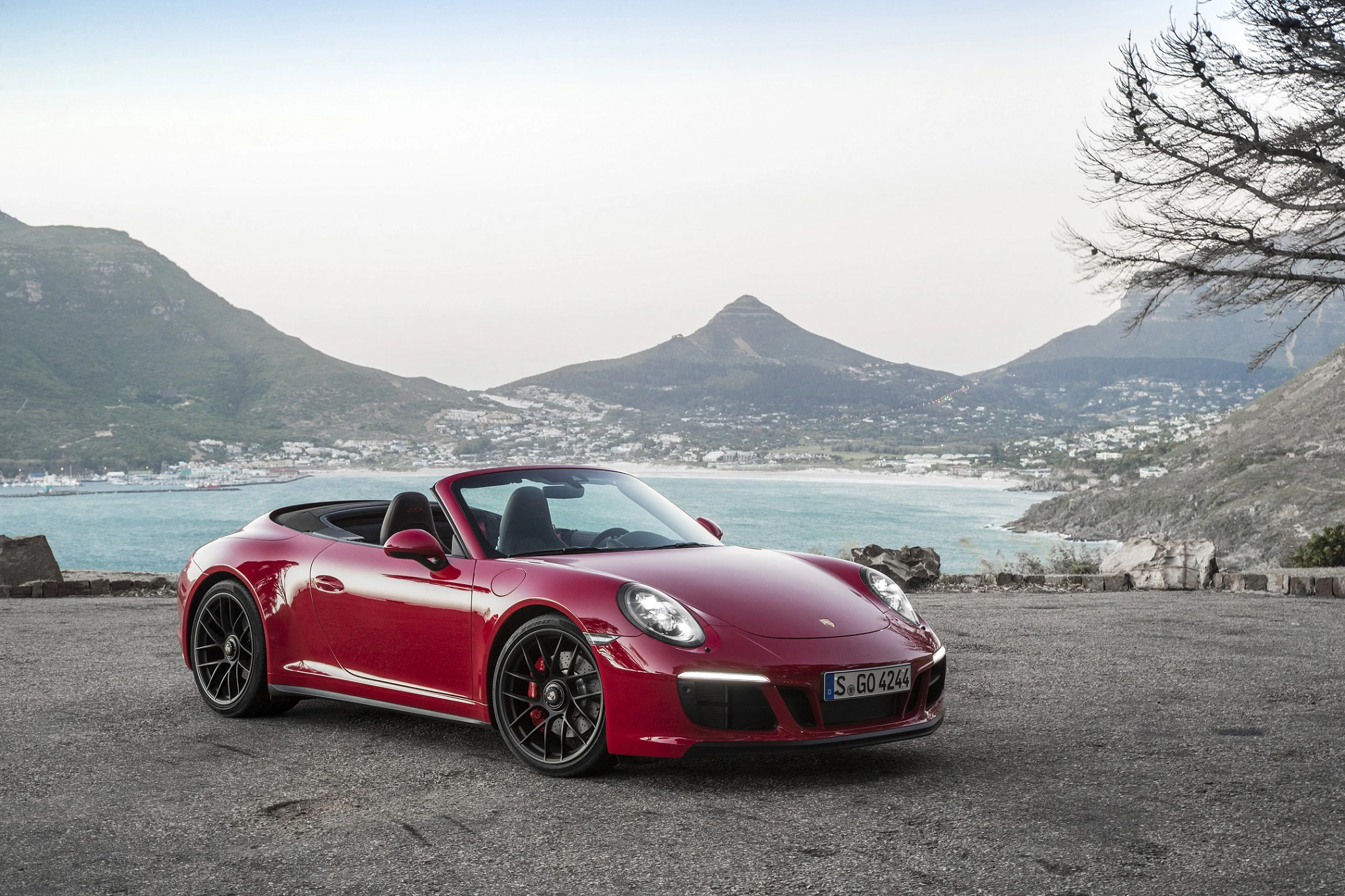 Roter Sportwagen-Cabrio geparkt vor einer Küstenlandschaft mit Bergen und Meer im Hintergrund. Klarer Himmel und wenige Bäume sichtbar.