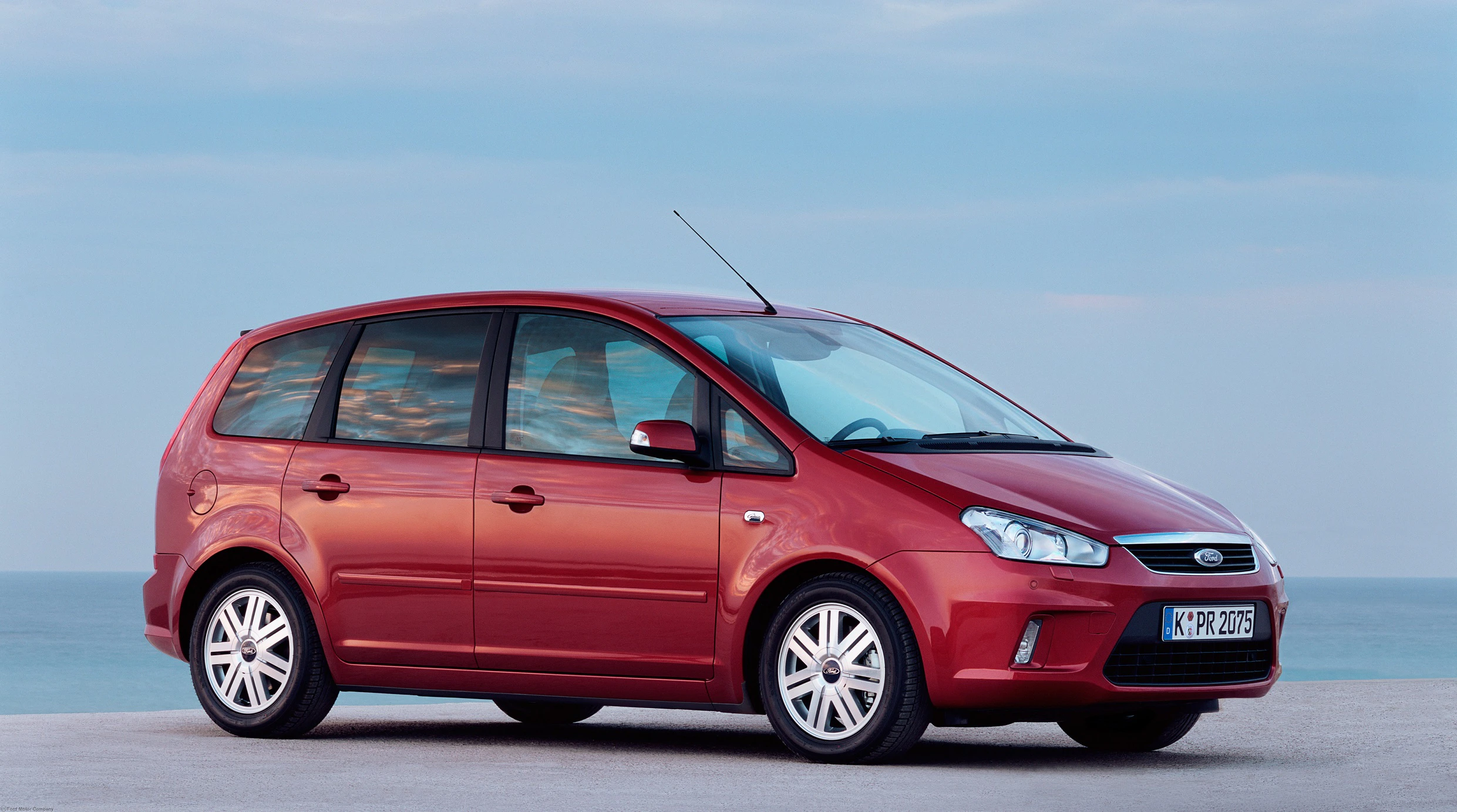 Rotes Ford C-Max Auto, geparkt auf einer Küstenstraße bei Sonnenuntergang, mit klarem Himmel und Meer im Hintergrund.