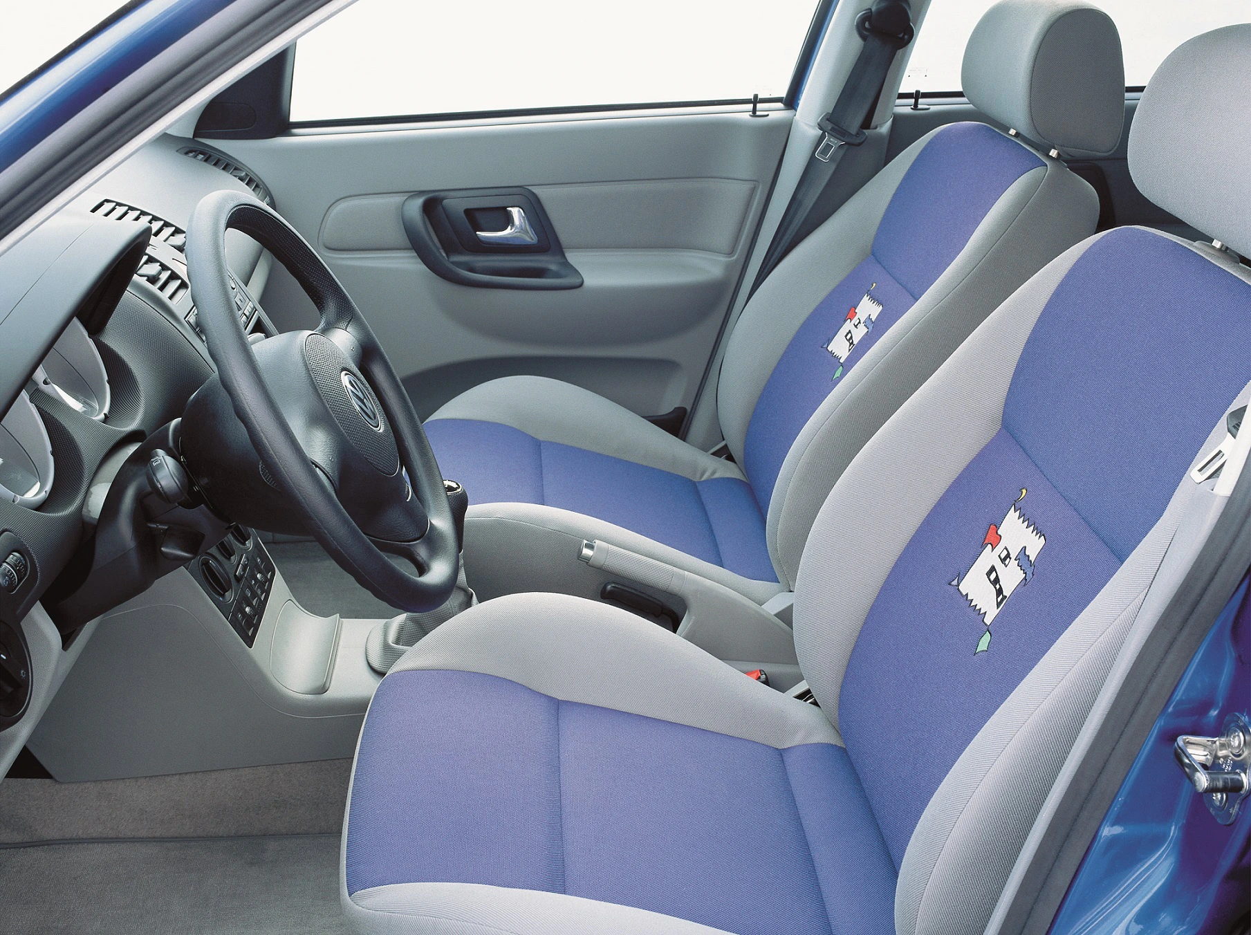 Innenraum eines Autos mit blau-grauen Sitzen und farbigem Muster. Lenkrad links, graue Türverkleidung mit Türgriff und Fensterheber sichtbar.