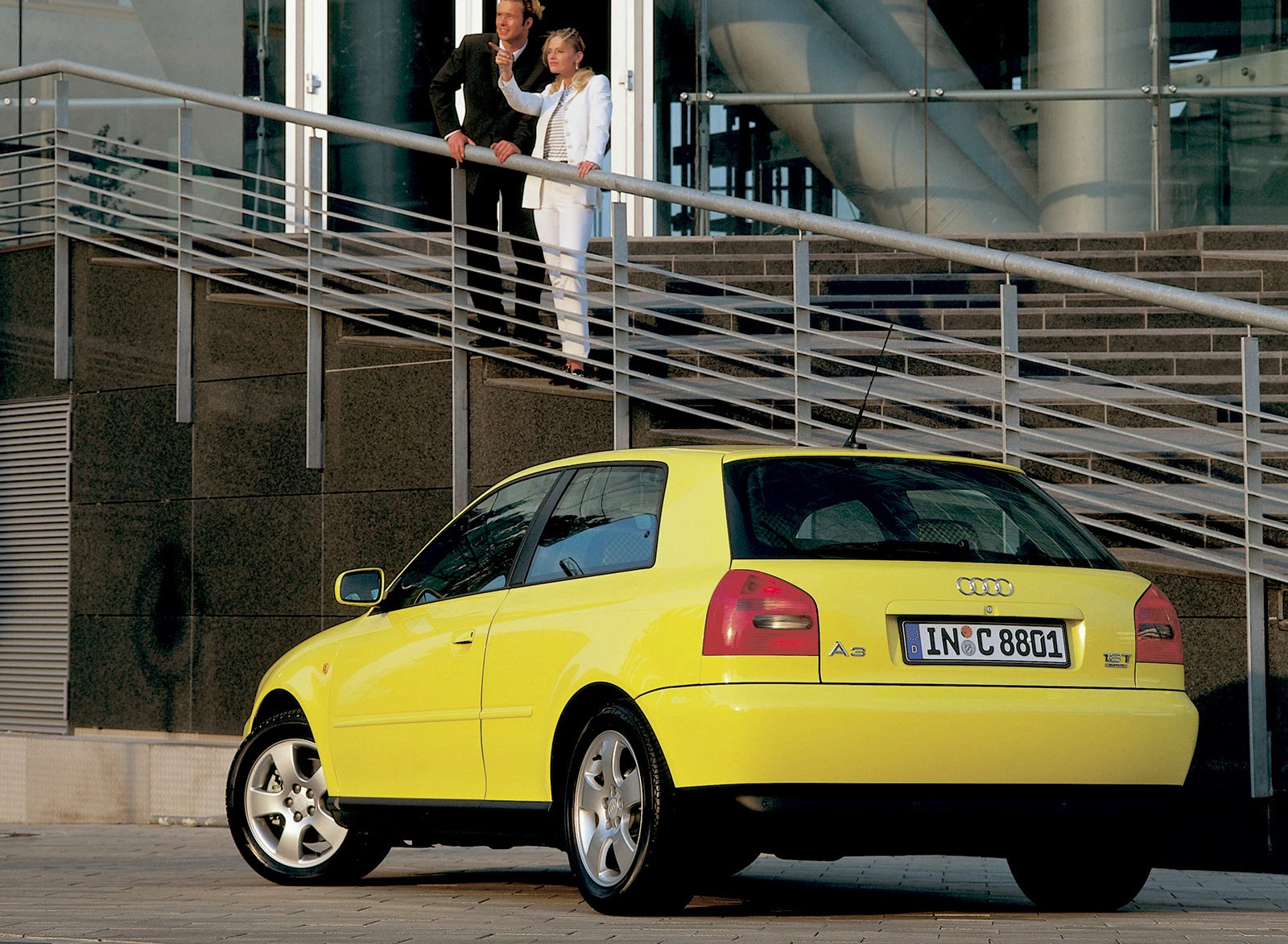 Gelber Audi A3 parkt vor einem modernen Gebäude. Zwei Personen in formeller Kleidung stehen auf einer Treppe und zeigen in die Ferne.