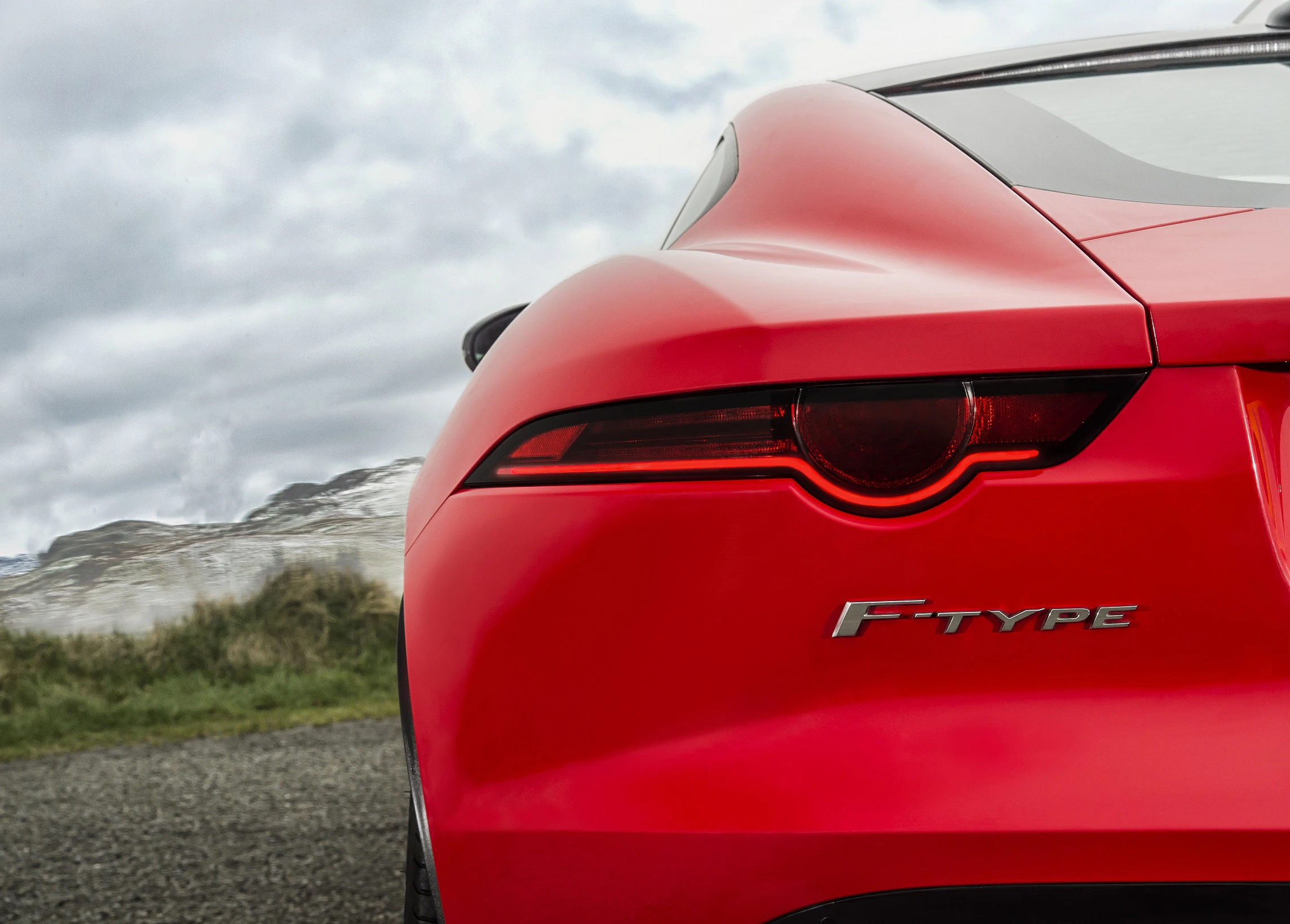 Nahaufnahme eines roten Sportwagen-Hecks mit dem Schriftzug "F-Type" vor einer Landschaft aus Hügeln und bewölktem Himmel.