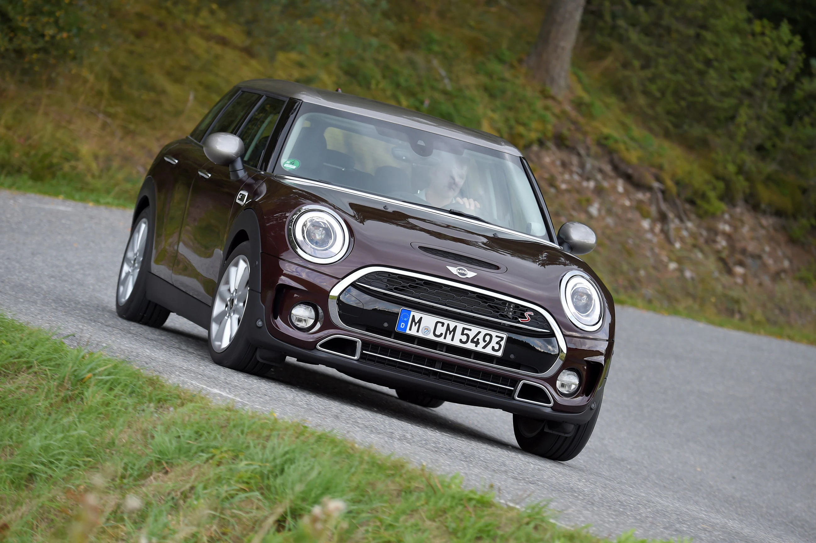 Ein dunkelroter Mini Cooper S fährt auf einer kurvigen Straße durch eine grüne, bewaldete Landschaft.