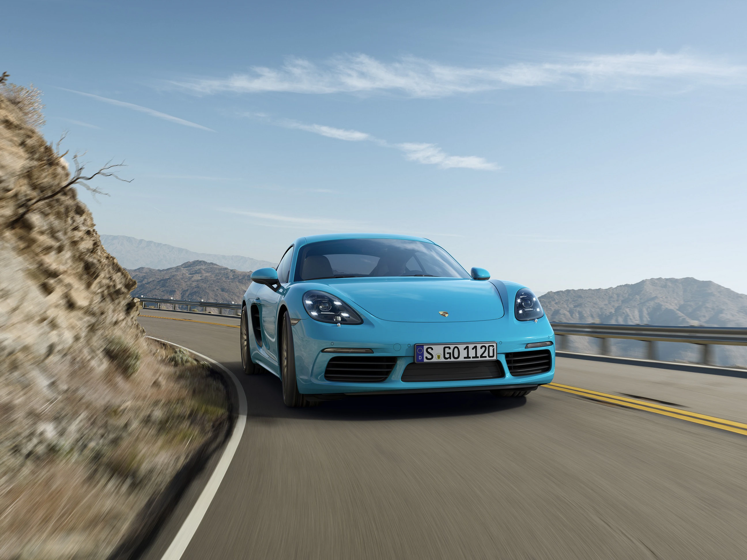 Ein blauer Sportwagen fährt auf einer kurvigen Bergstraße bei klarem Himmel. Im Hintergrund sind Felsen und Berge zu sehen.