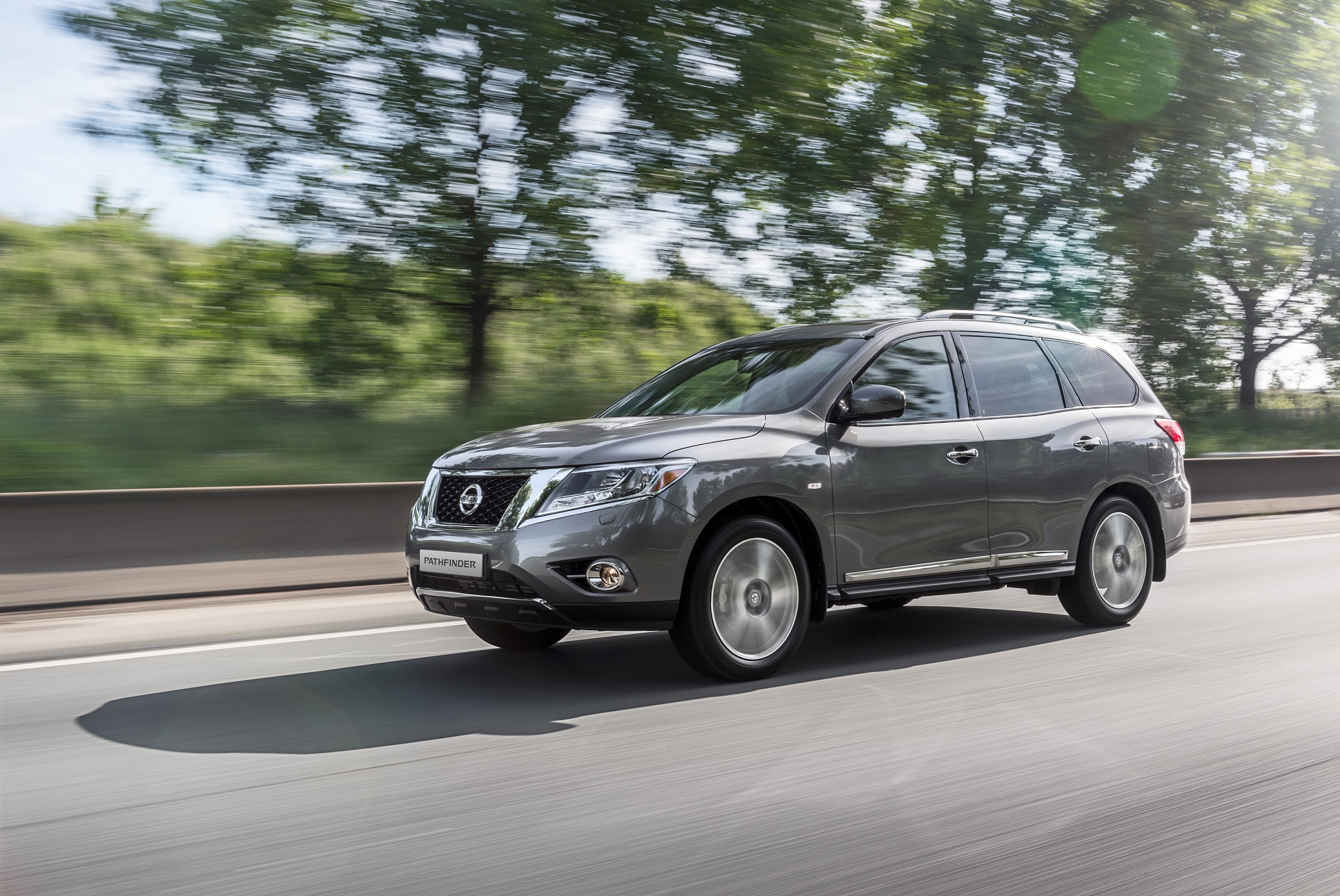 Grauer Nissan-SUV fährt auf einer Landstraße, umgeben von grünen Bäumen und leicht bewölktem Himmel.