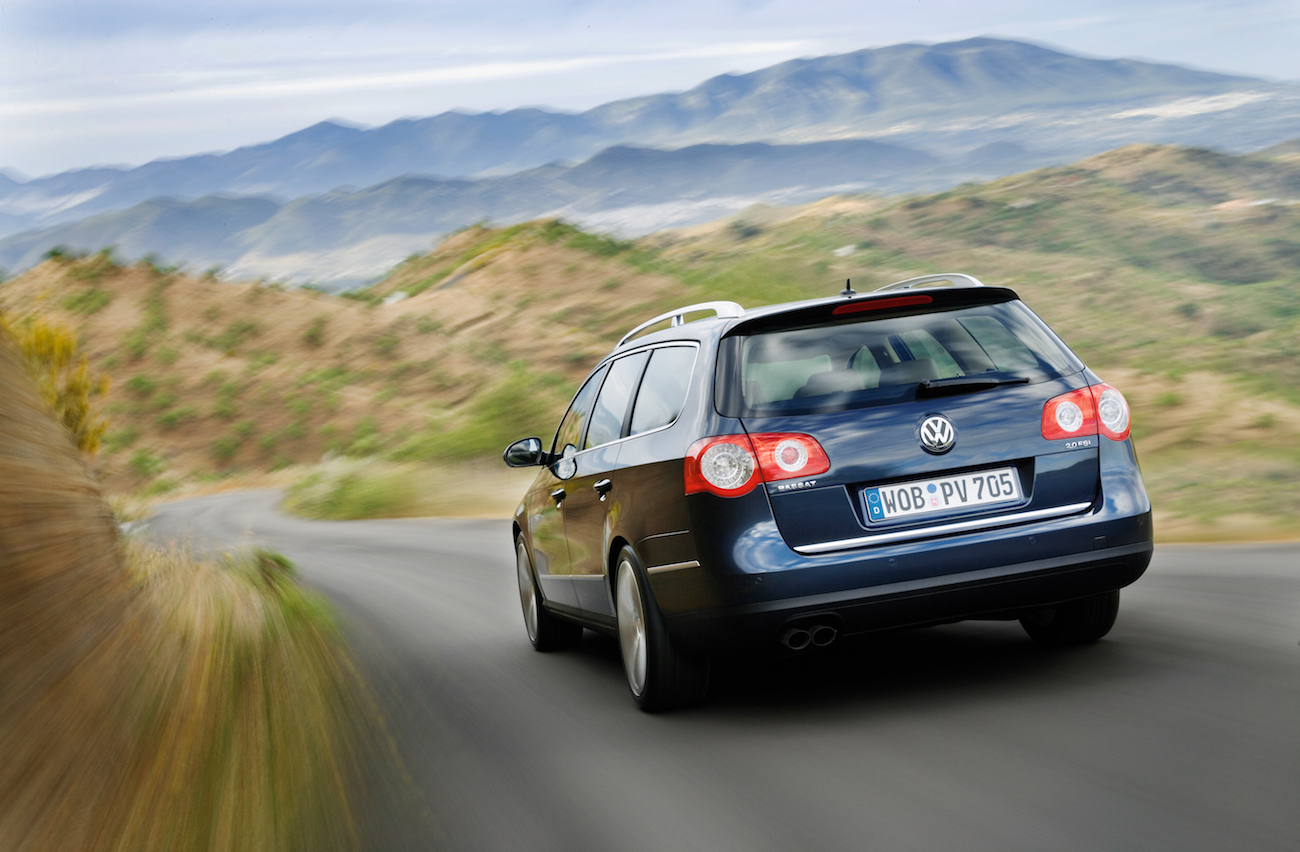 Silberner Volkswagen Kombi fährt auf einer kurvigen Straße durch eine hügelige Landschaft mit Bergen im Hintergrund.