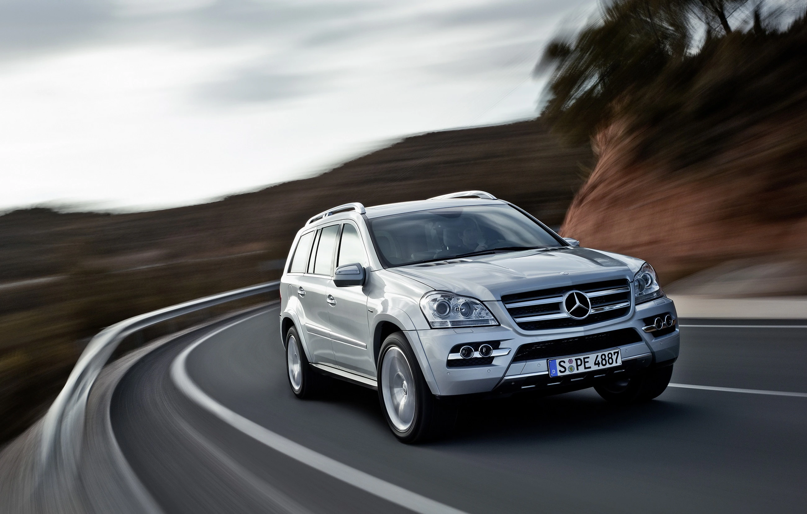 Silberner Mercedes-SUV fährt schnell auf kurviger Landstraße. Hintergrund verschwommen mit Naturkulisse.